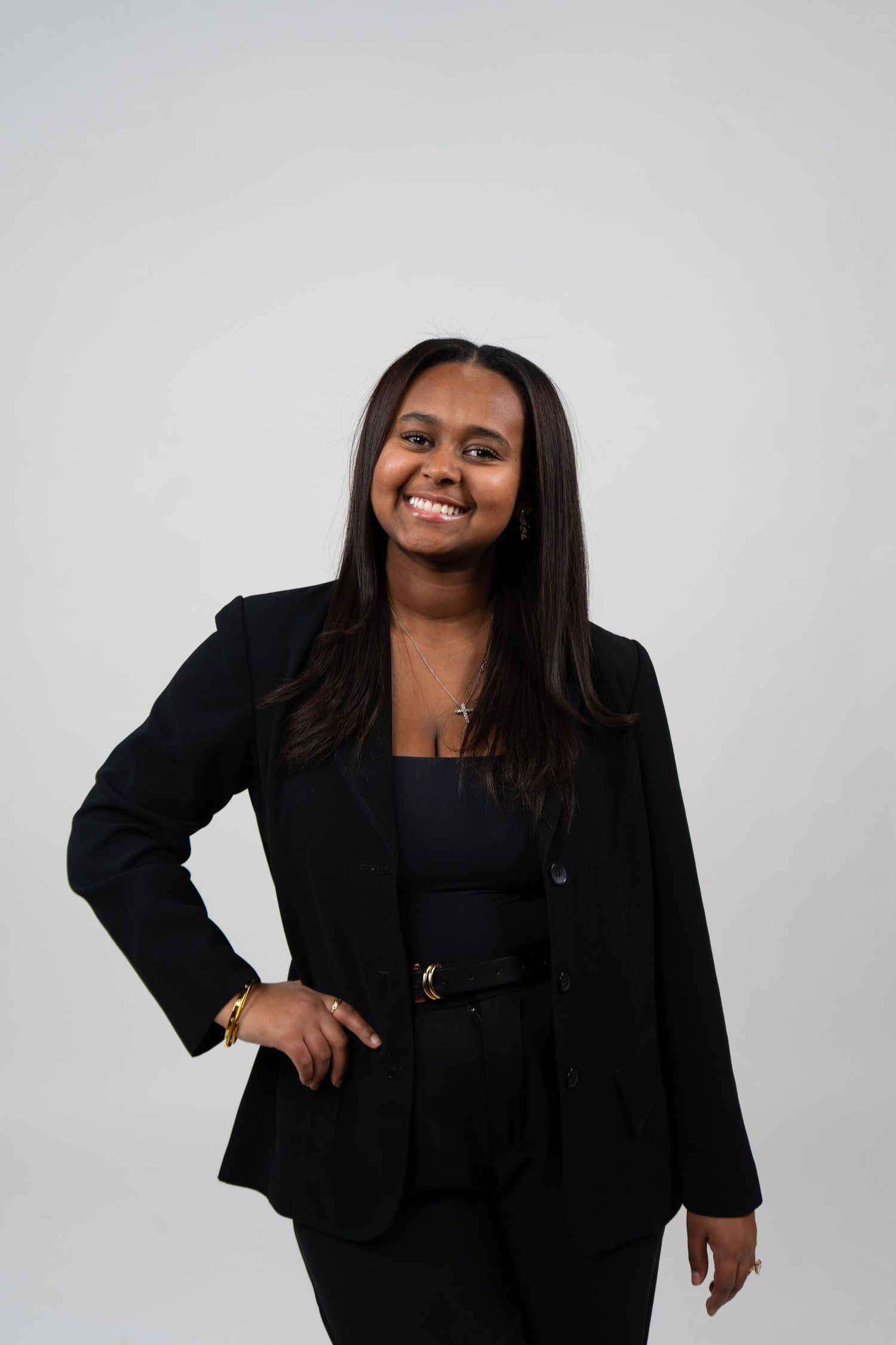 Young professional woman in black blazer and pants smiling confidently against a light gray background.
