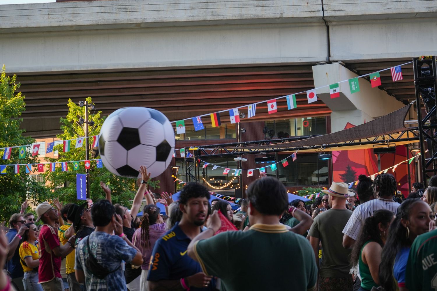 Crowd at an outdoor soccer festival with a giant inflatable soccer ball, colorful international flags, and an overpass backdrop.
