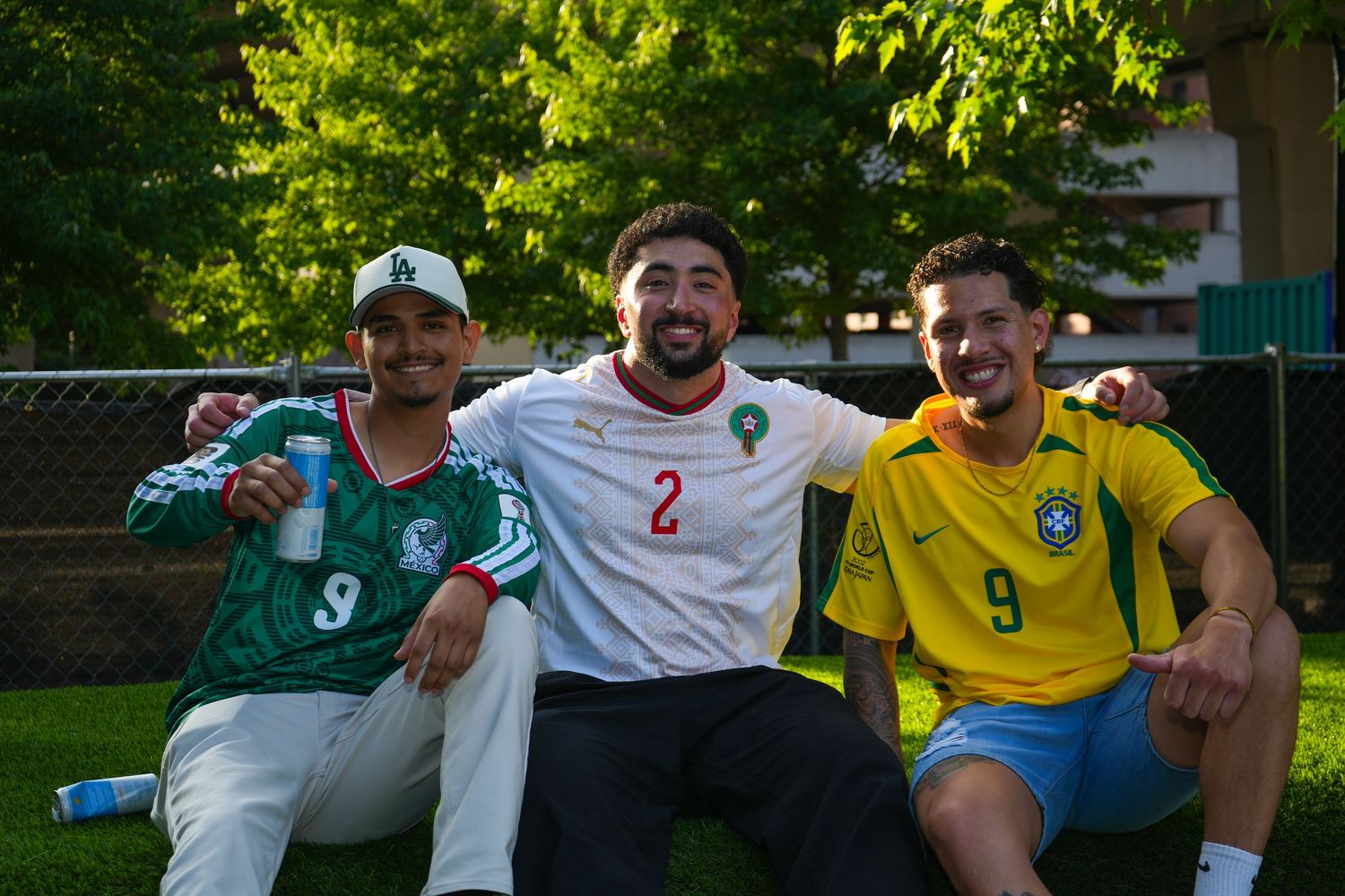 Three friends posing outdoors in Mexico, Morocco, and Brazil soccer jerseys, smiling at a sports event.