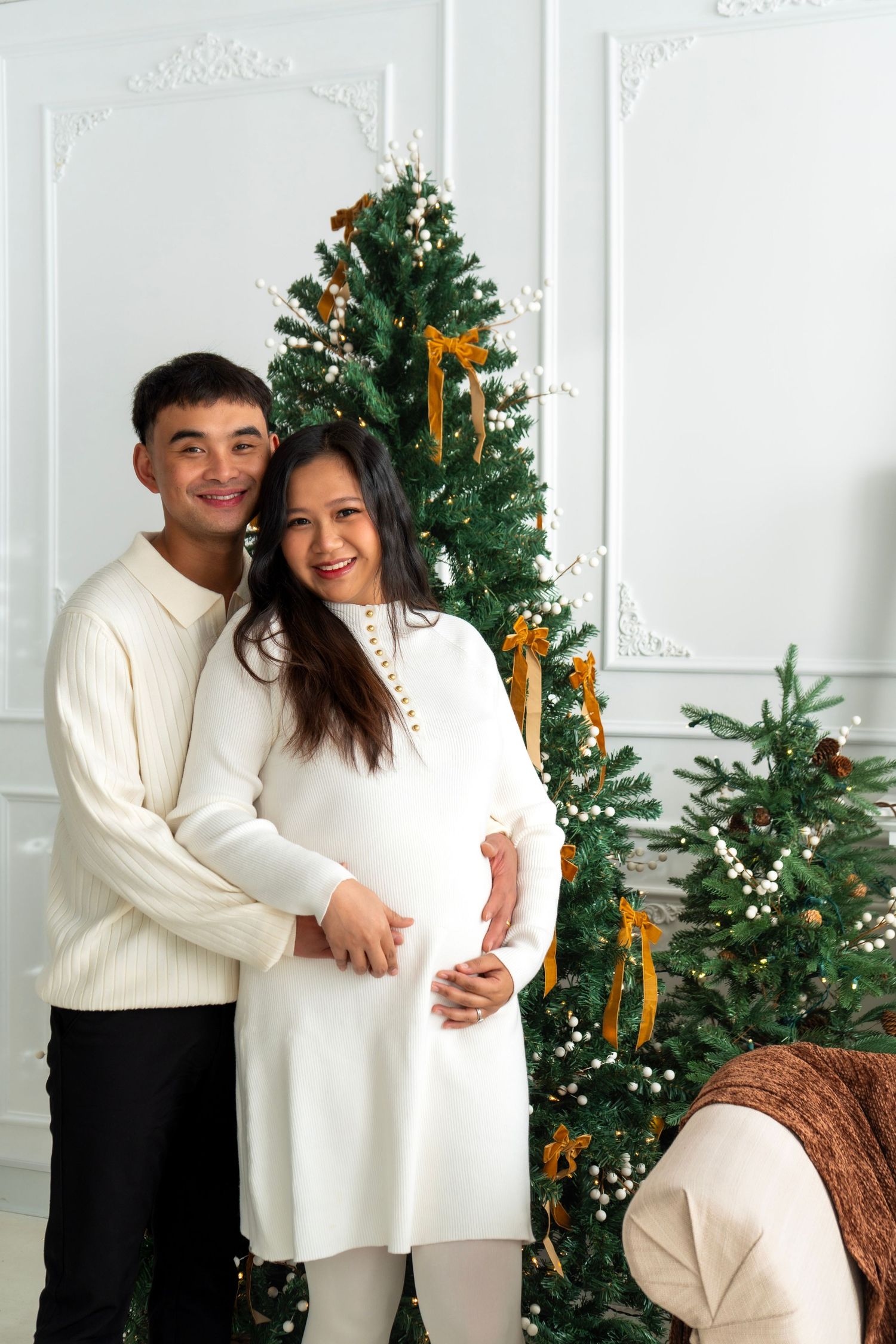 Happy couple in white outfits embracing by decorated Christmas tree, pregnant woman cradling baby bump in festive room.