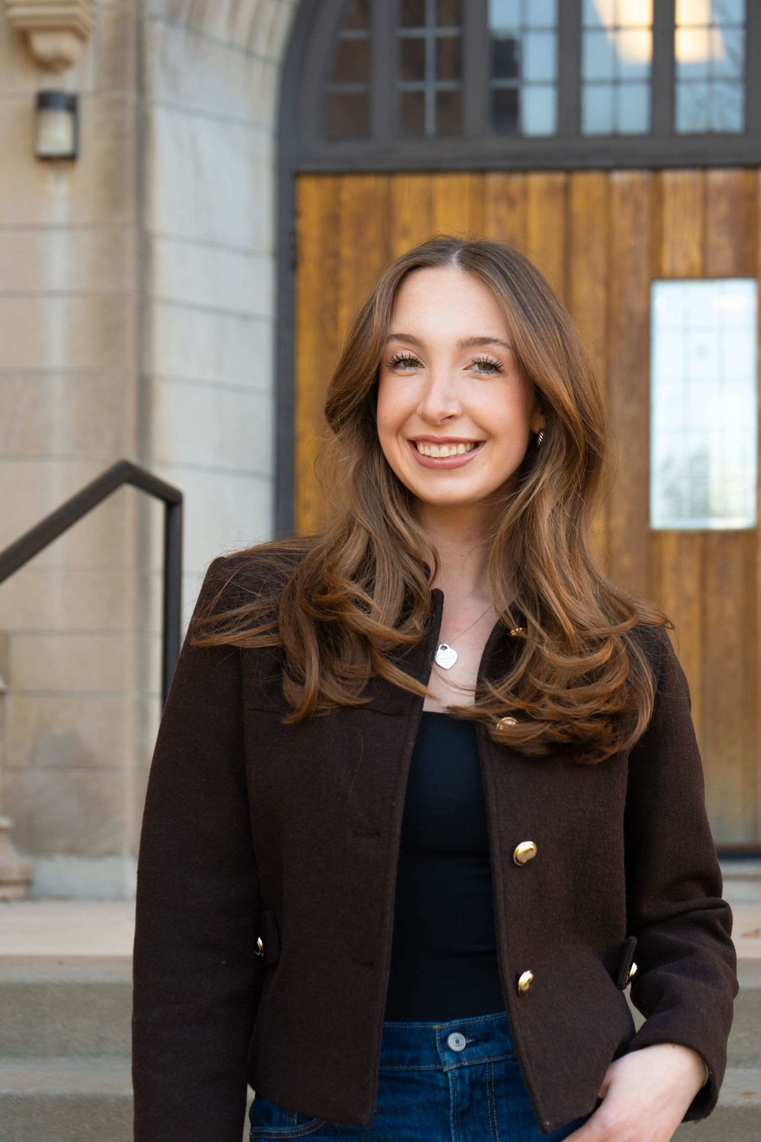 Young woman with long brown hair smiling in dark blazer with gold buttons, standing outside a stone building.