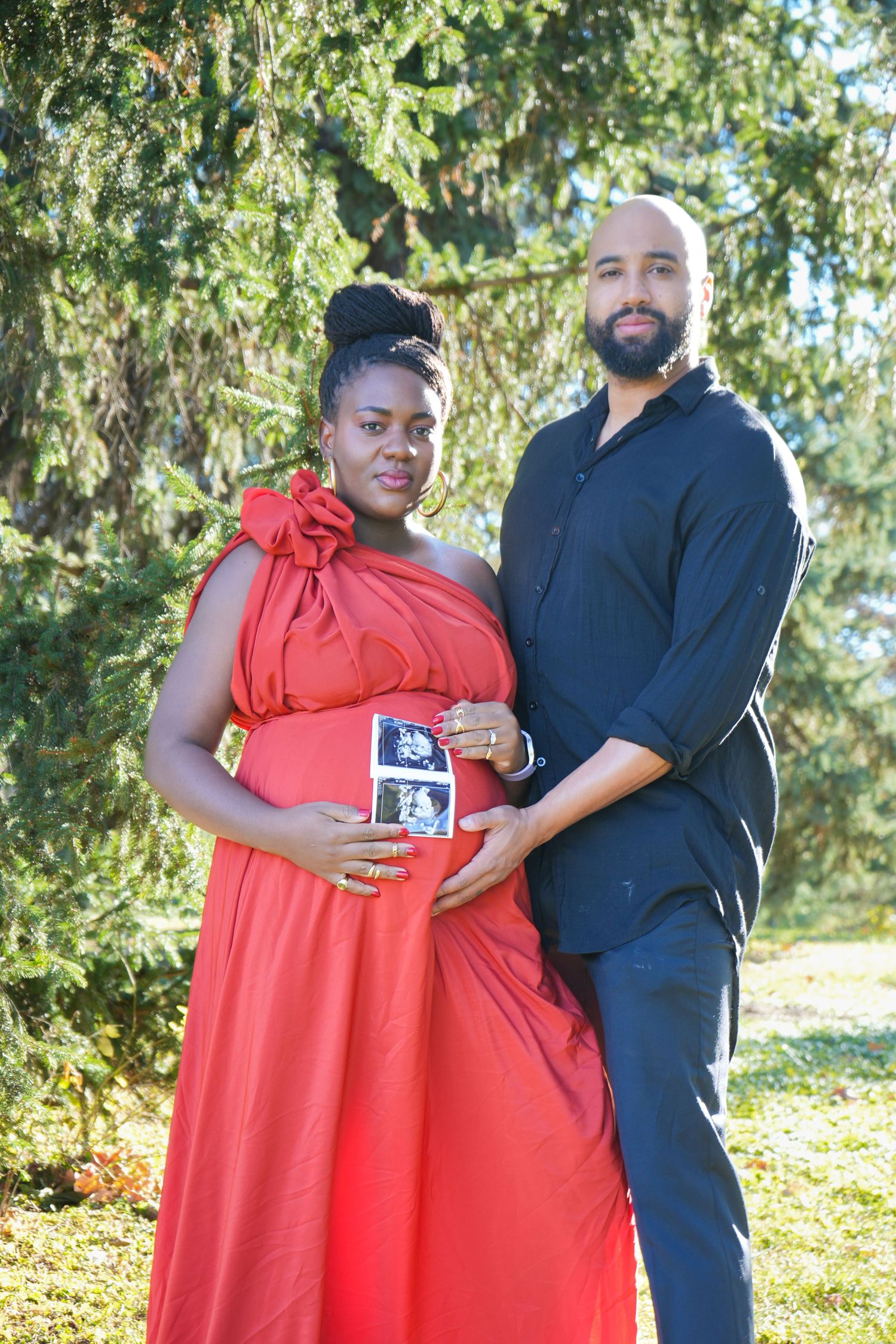 Pregnant couple in park, woman in red dress holding ultrasound photos, man in black shirt, maternity announcement photo.