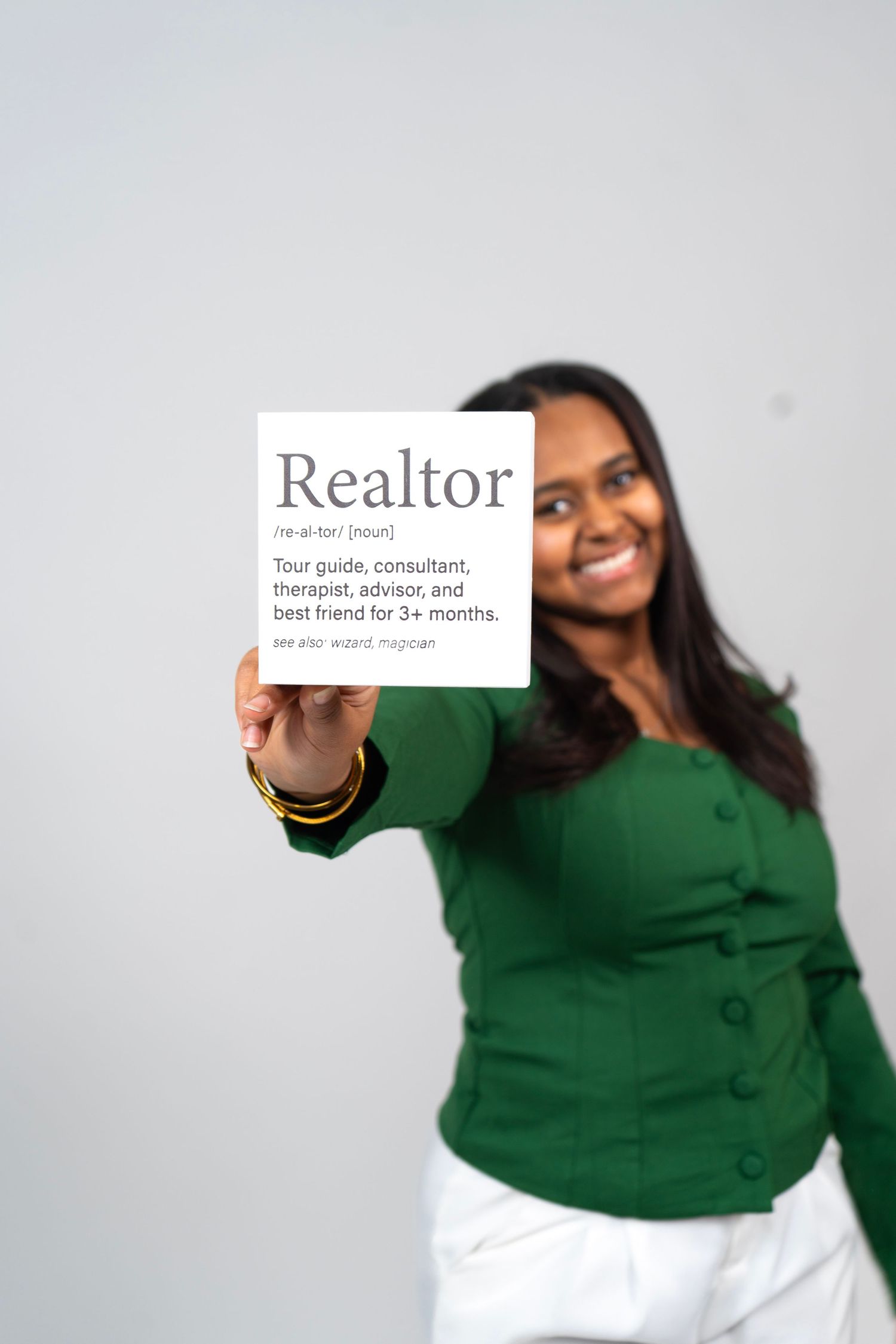 Smiling woman in green sweater holding a Realtor definition card describing tour guide, consultant, therapist, and best friend.