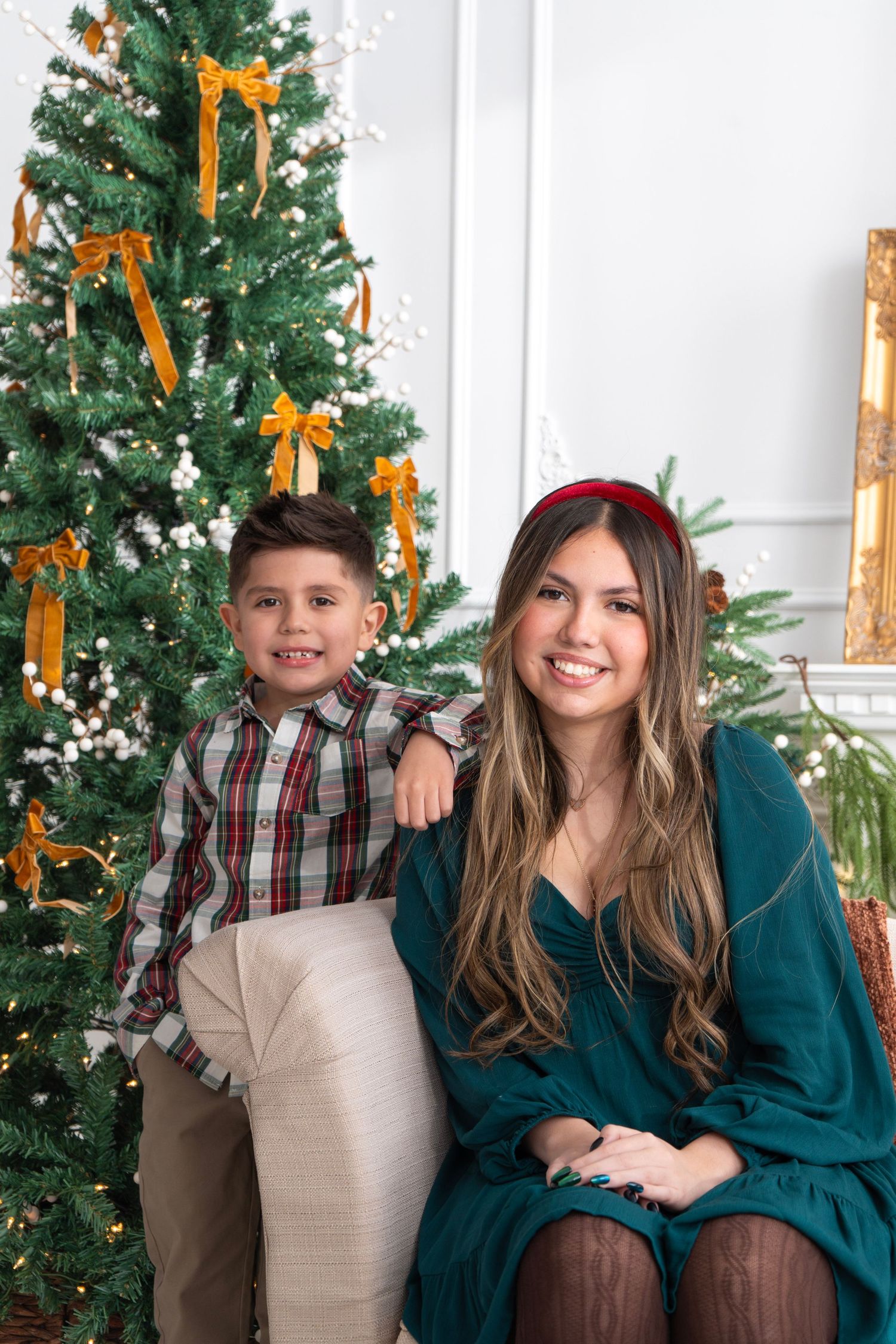 A young boy and woman smile together in front of a decorated Christmas tree with gold ribbons indoors.