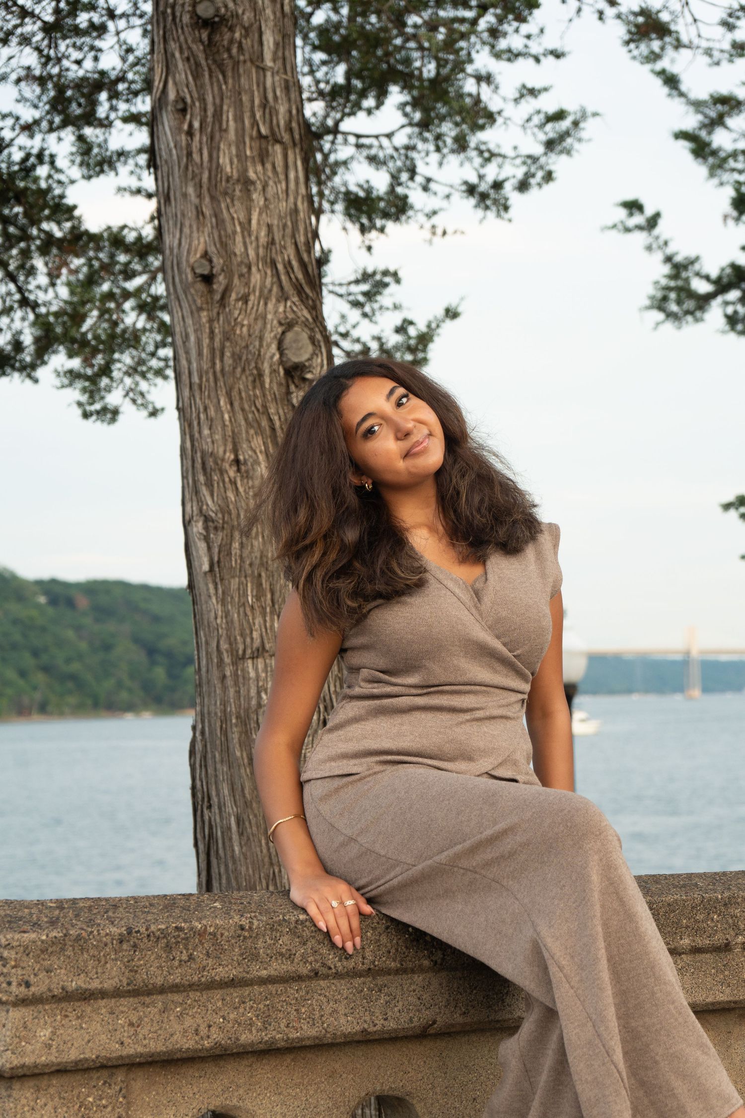 Young woman in gray dress sitting by a tree overlooking a river, smiling warmly in a serene outdoor setting.