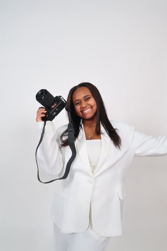 Young woman in white suit smiling and holding a black DSLR camera against a light gray background.
