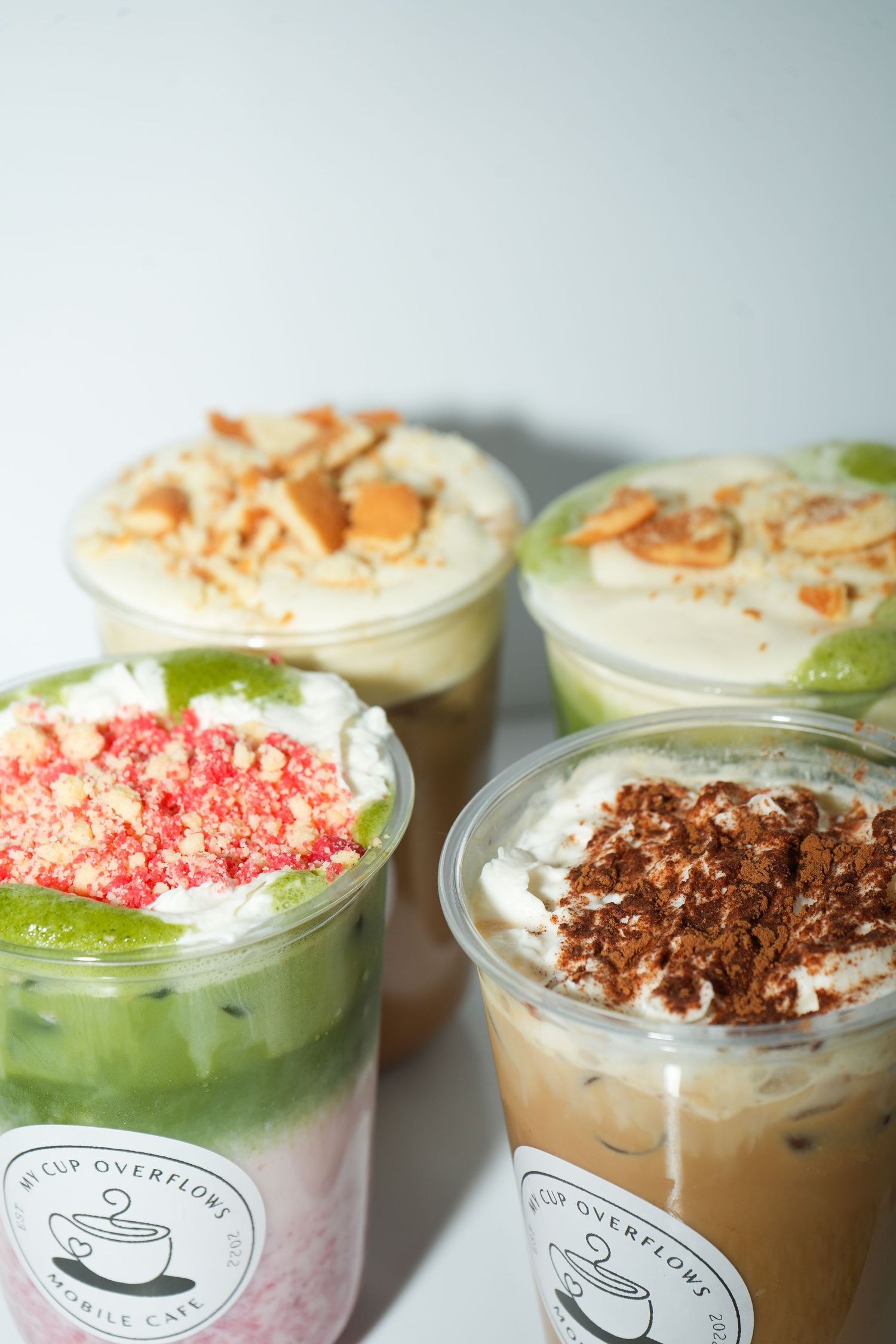 Four colorful layered dessert drinks in plastic cups topped with chocolate, almonds, and strawberry crumbles at Cup Overflows Cafe.