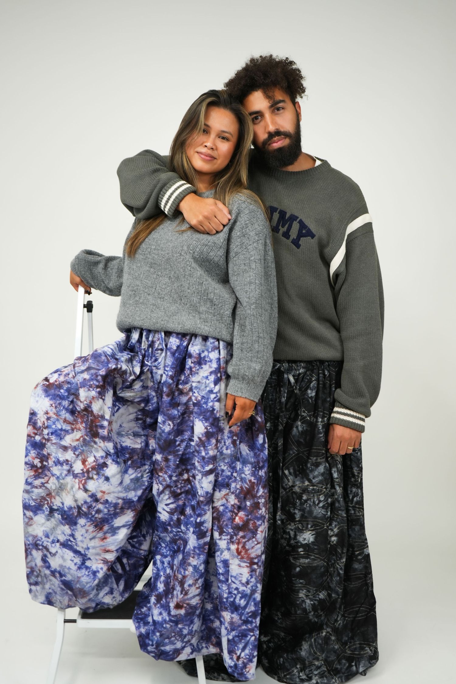 A man and woman pose together wearing matching gray sweatshirts and floral tie-dye maxi skirts in a studio.