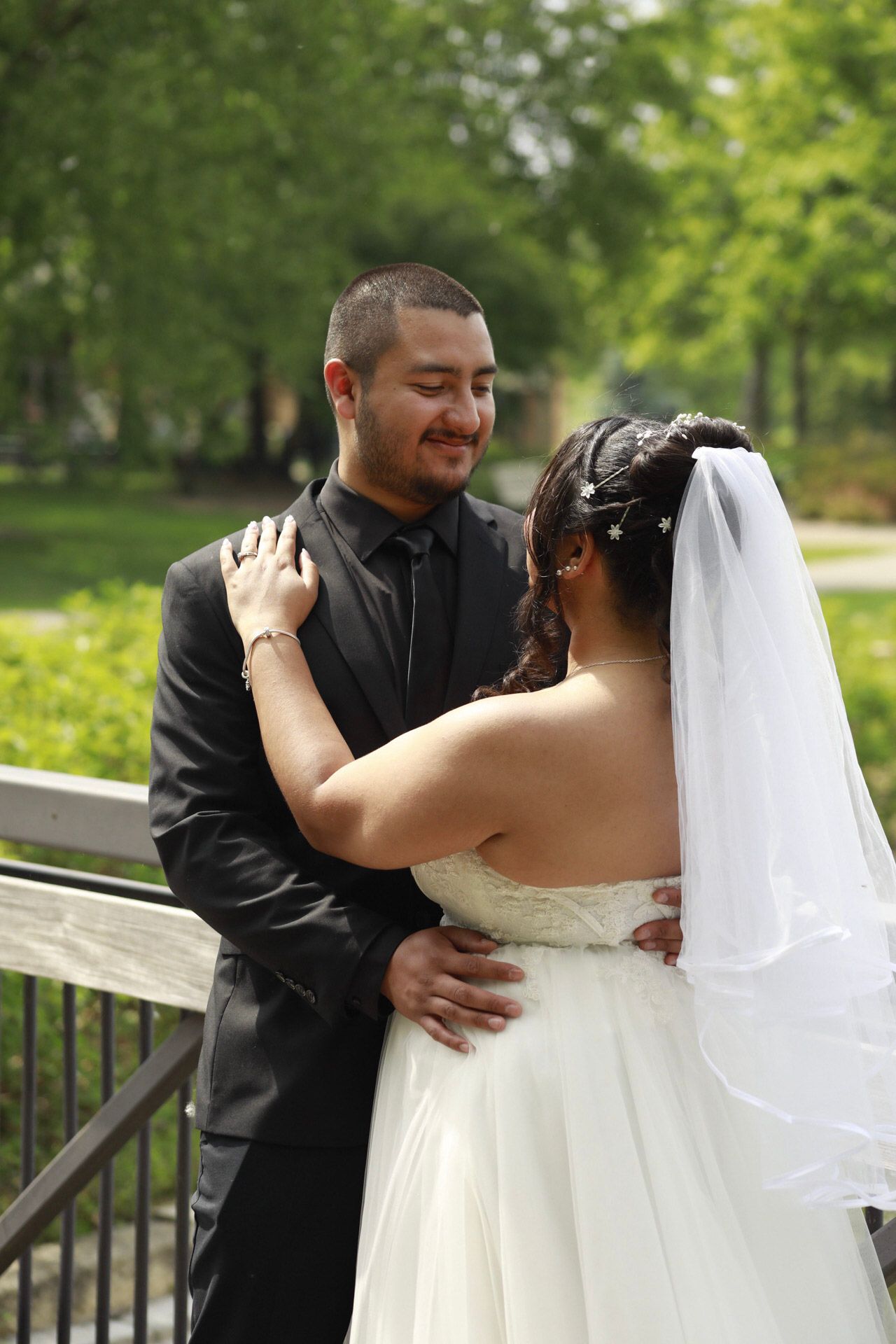 A groom in a black suit smiles at his bride in a white wedding gown and veil, embracing outdoors in a park.