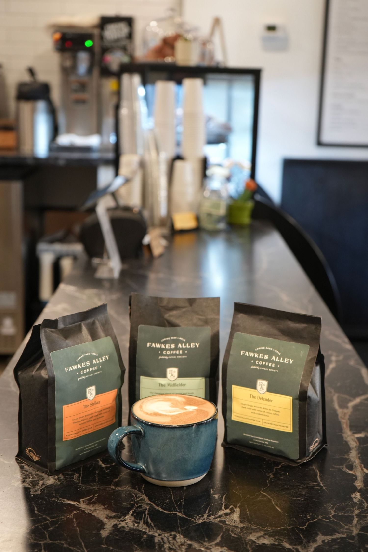 Three Fawker Alley Coffee bags displayed around a blue latte mug on a dark counter in a modern cafe.
