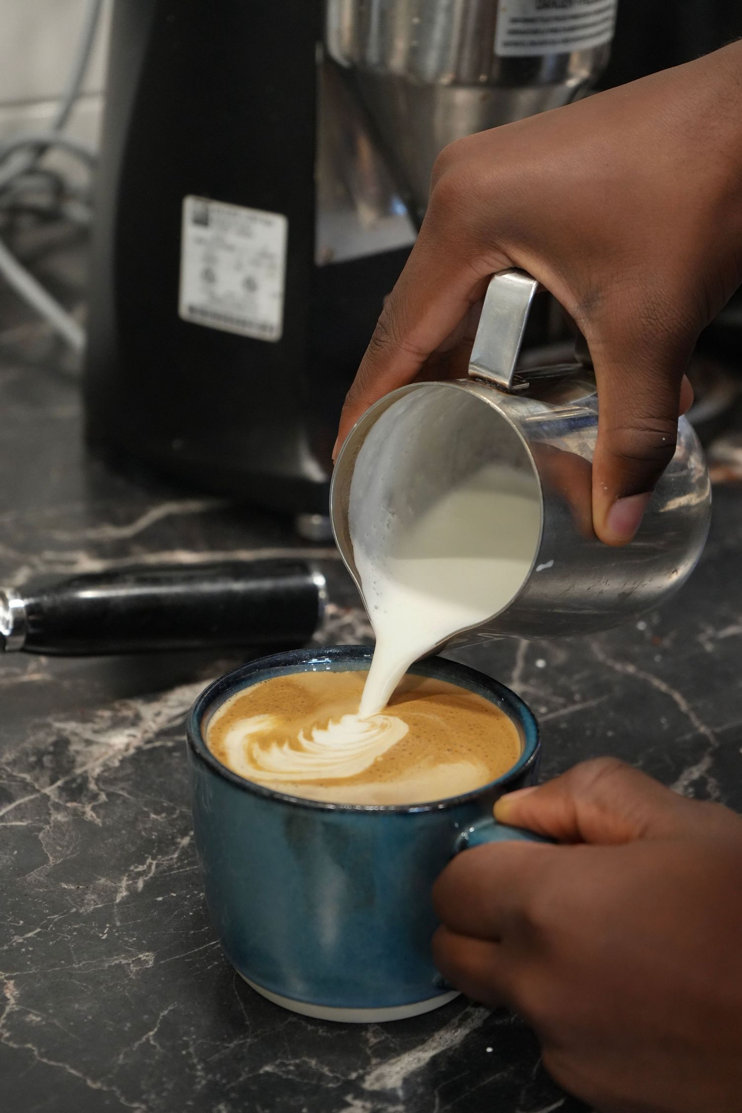 Barista pouring steamed milk into a blue ceramic cup creating latte art on a marble counter.