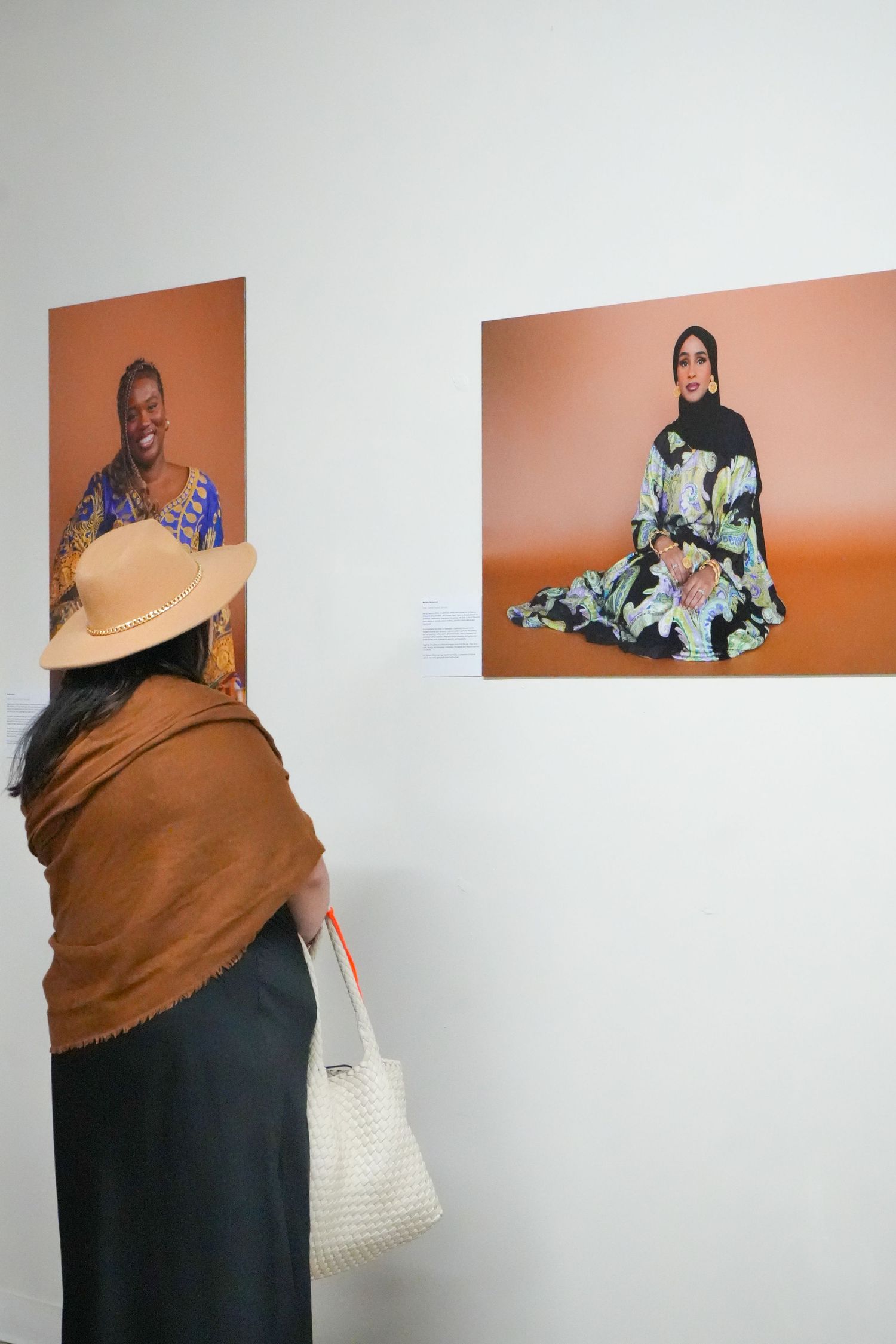 Woman in tan hat and brown shawl views multicultural portrait photographs at an art gallery exhibition.