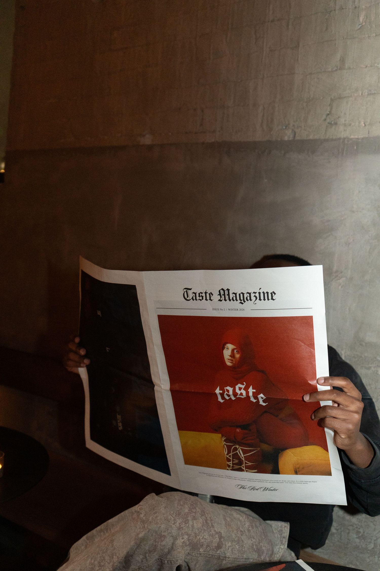 Person holding Taste Magazine open, featuring a red cover with a model wearing a hoodie, in a dimly lit setting.