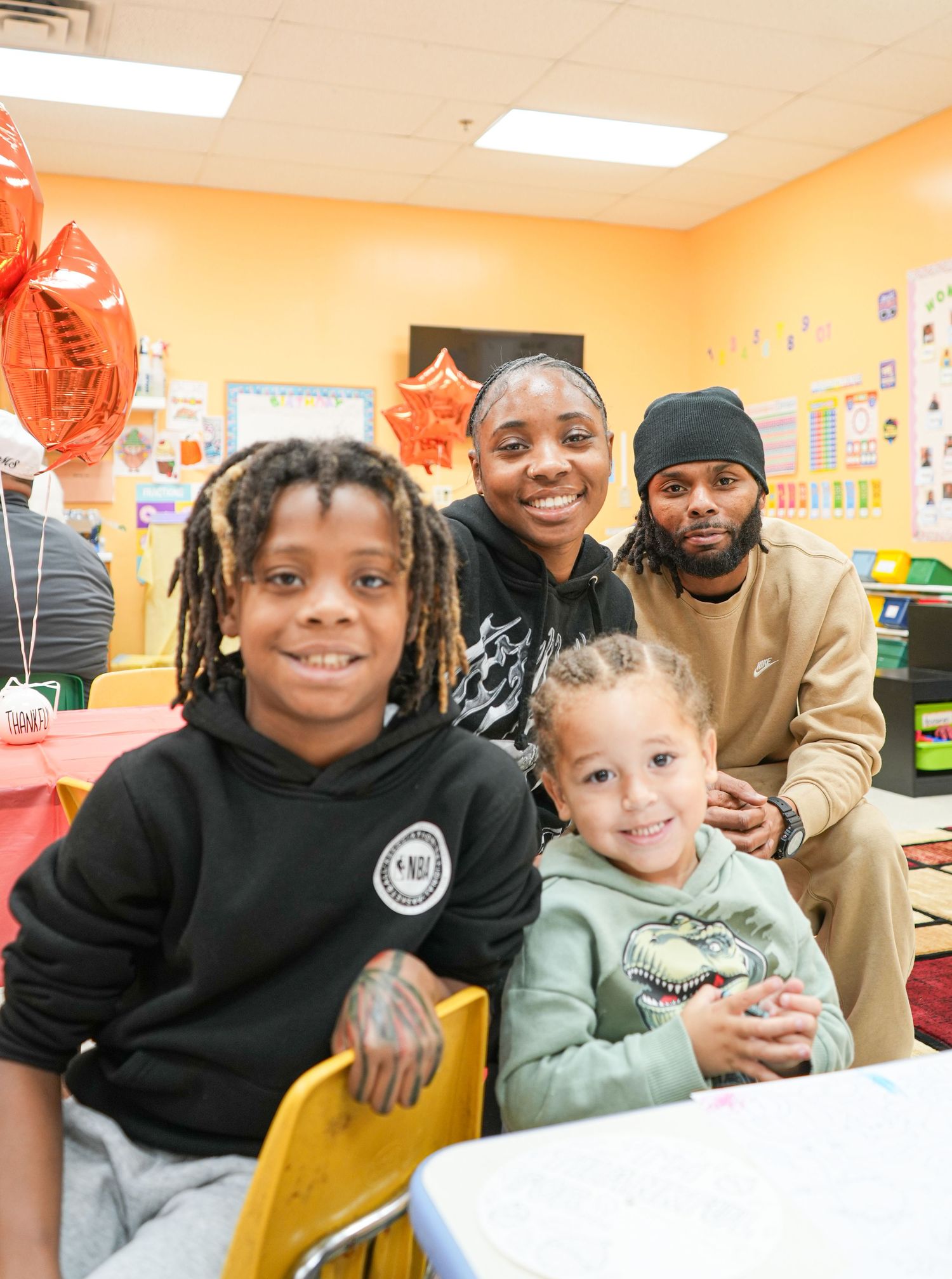 Family of four smiling together at a colorful classroom party with red star balloons in the background.