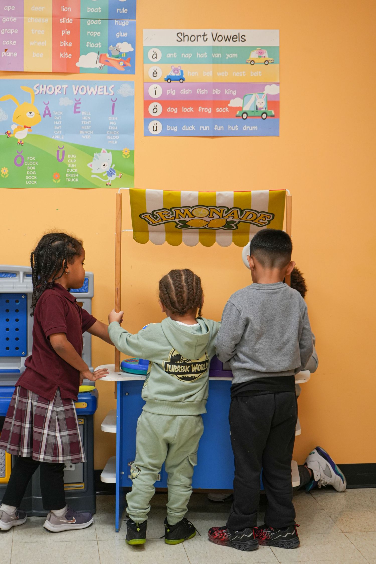 Three children play at a lemonade stand in a colorful classroom with short vowels educational posters on the wall.