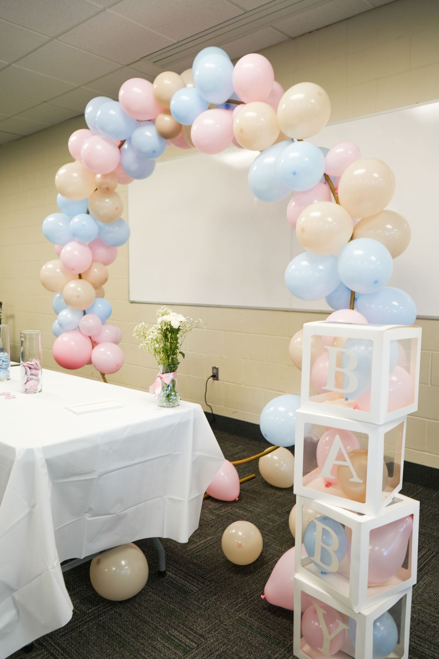Baby shower balloon arch in pink, blue, and cream with transparent BABY letter blocks and white decorated table.