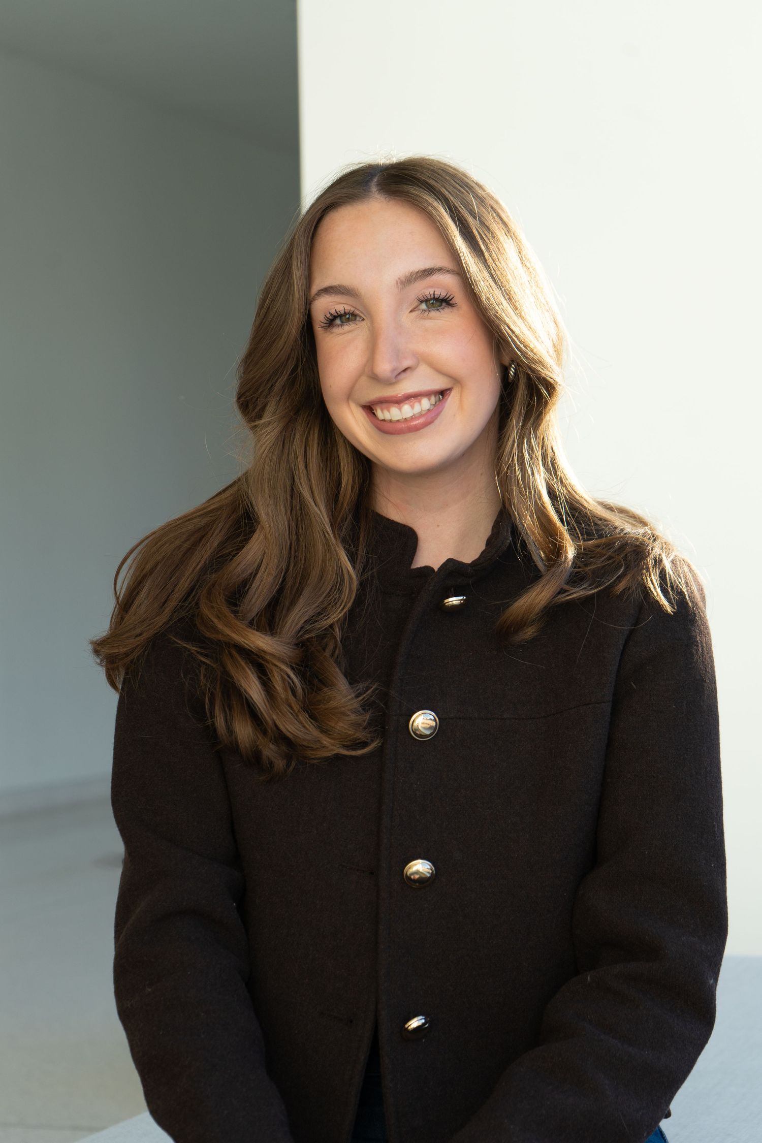 Smiling young woman with long brown hair wearing a black blazer with gold buttons, posing indoors.