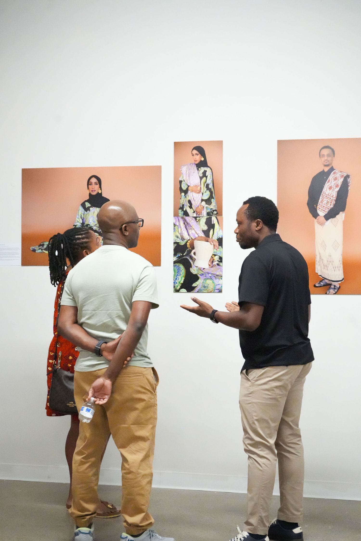 Three people discussing artwork at a gallery exhibition featuring portrait photographs on an orange background.