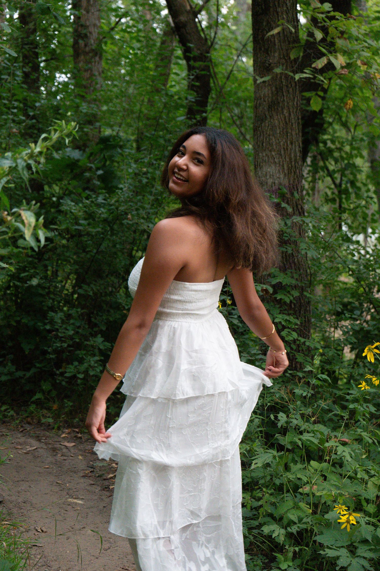 Young woman in white strapless tiered dress smiling over shoulder on forest path surrounded by lush green trees.