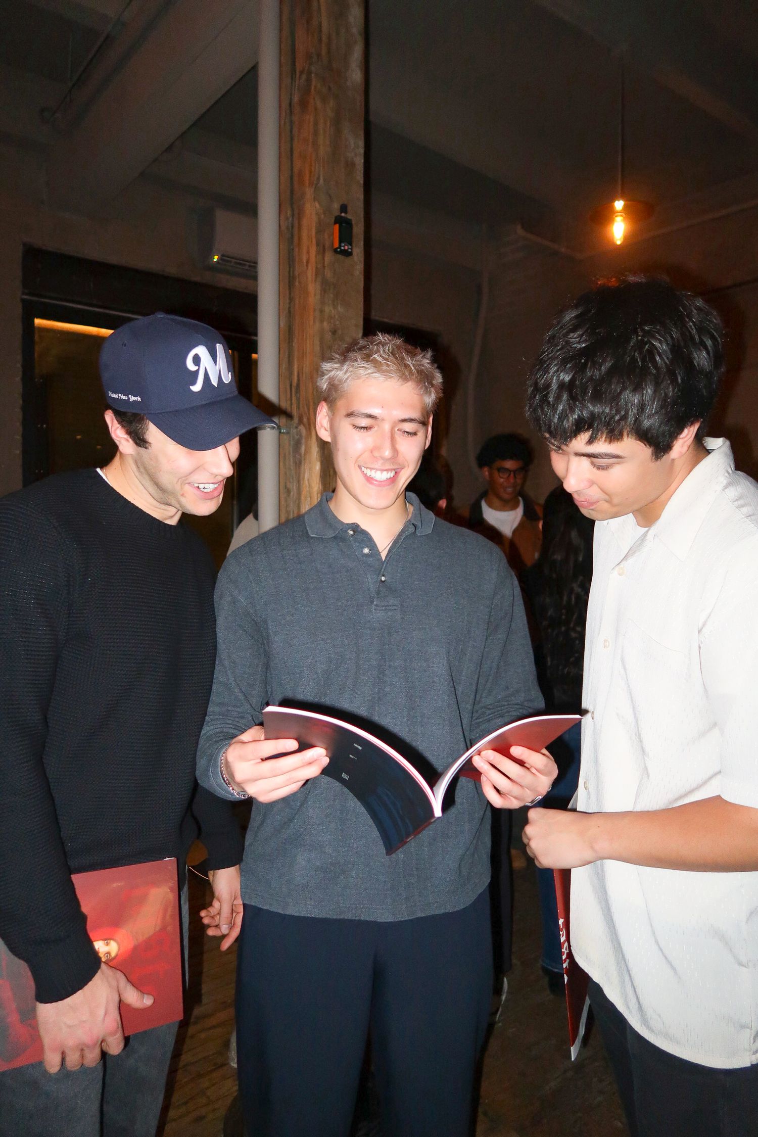 Three young men smiling and reading a magazine together indoors at a casual evening event.