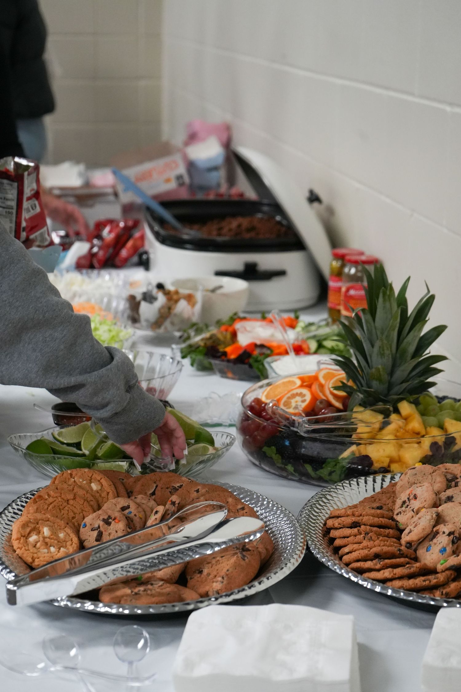Buffet table with cookie platters, fresh fruit display with pineapple, and assorted party foods at a catered event.