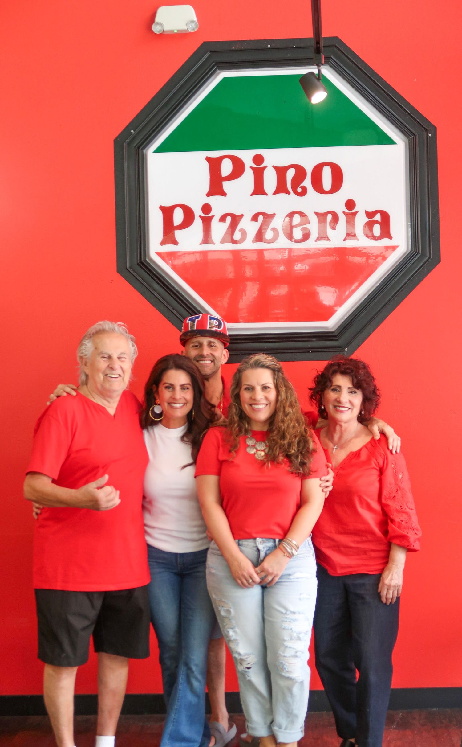 Five people smiling in front of Pino Pizzeria sign on red wall.