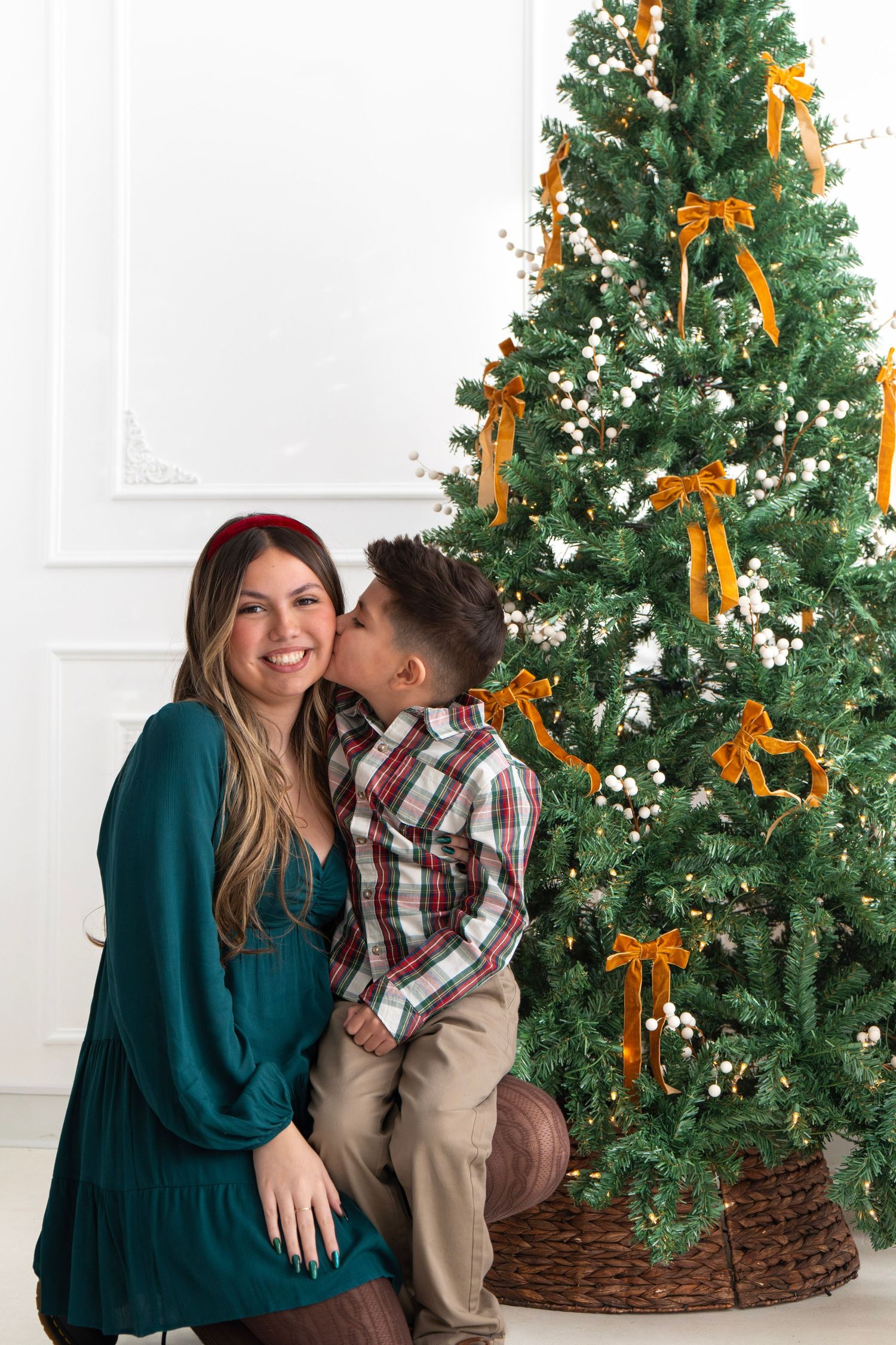 A young boy kisses his smiling mother on the cheek beside a decorated Christmas tree with gold bows.
