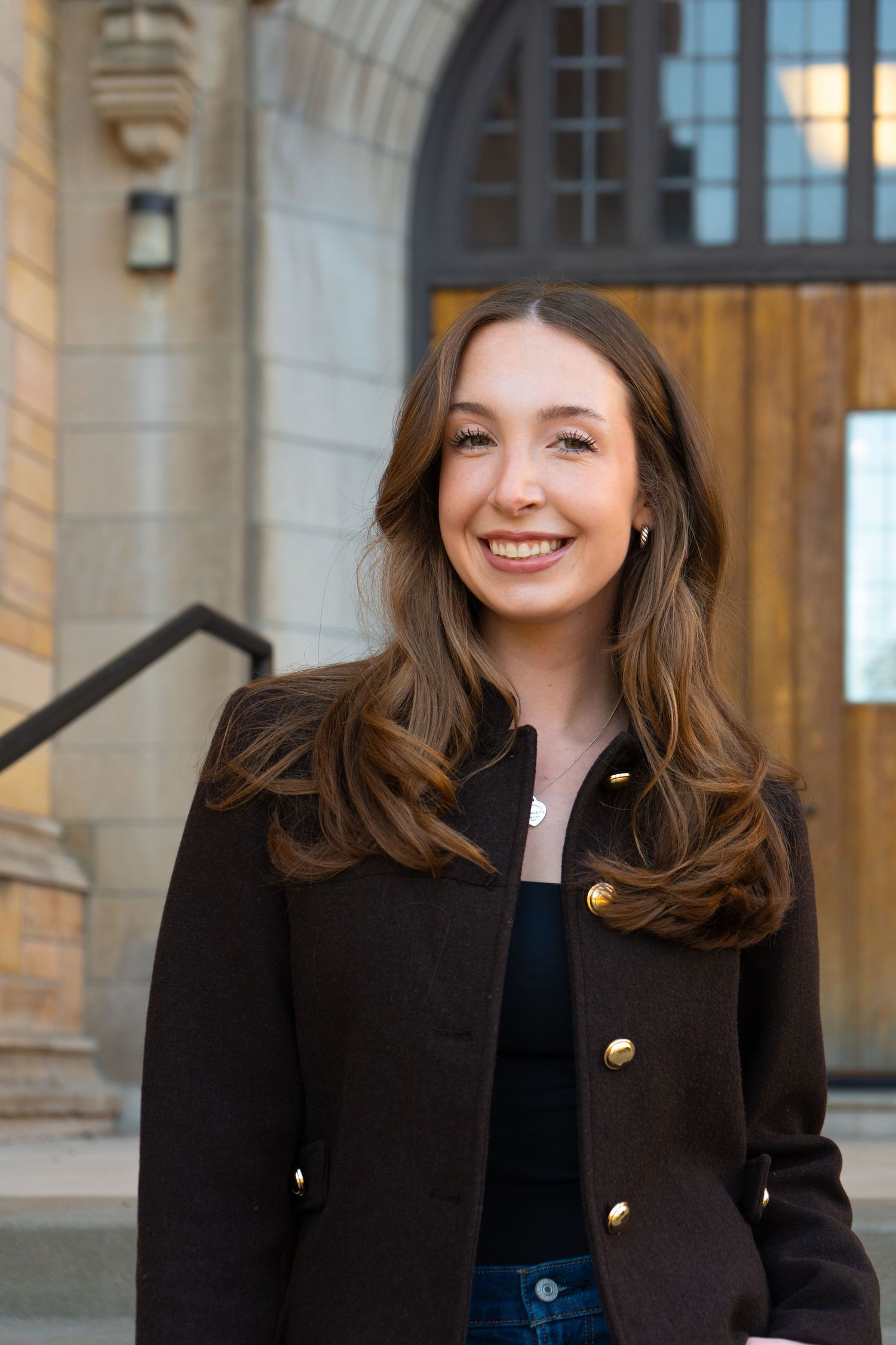 Smiling young woman in a black blazer with gold buttons standing in front of a stone building entrance.