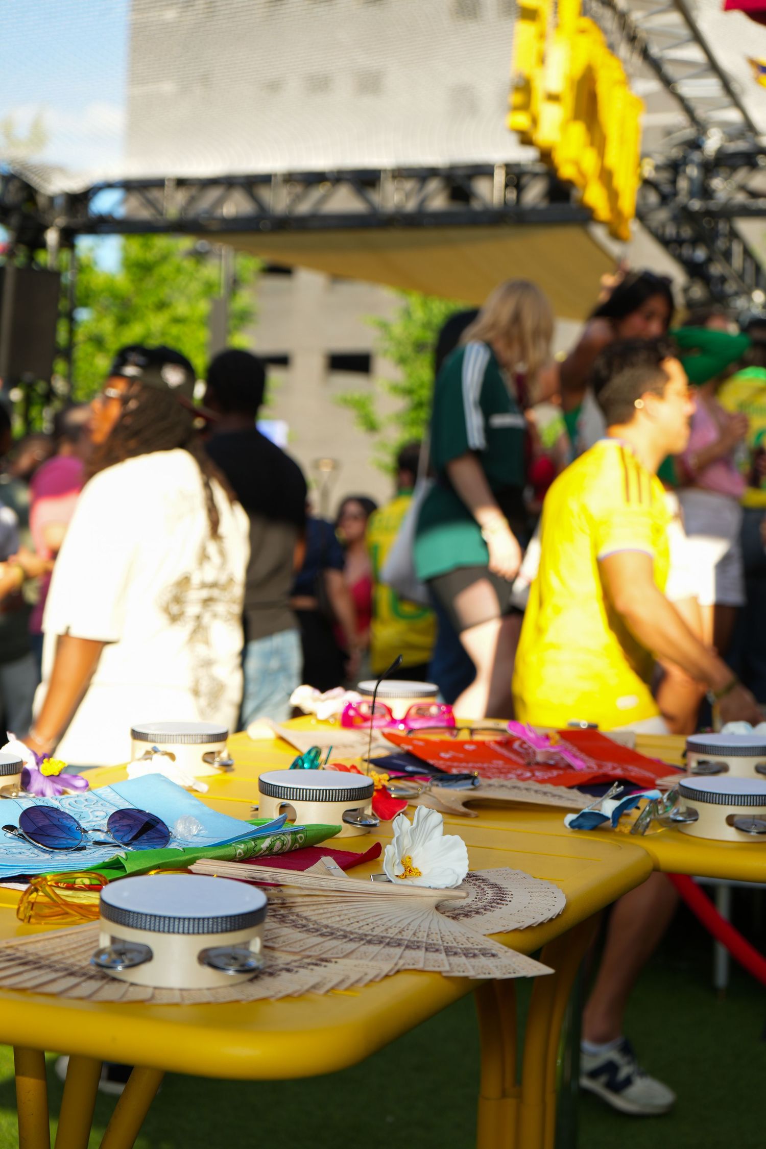 Colorful outdoor festival table with accessories and sunglasses, crowds dancing near a stage in the background.