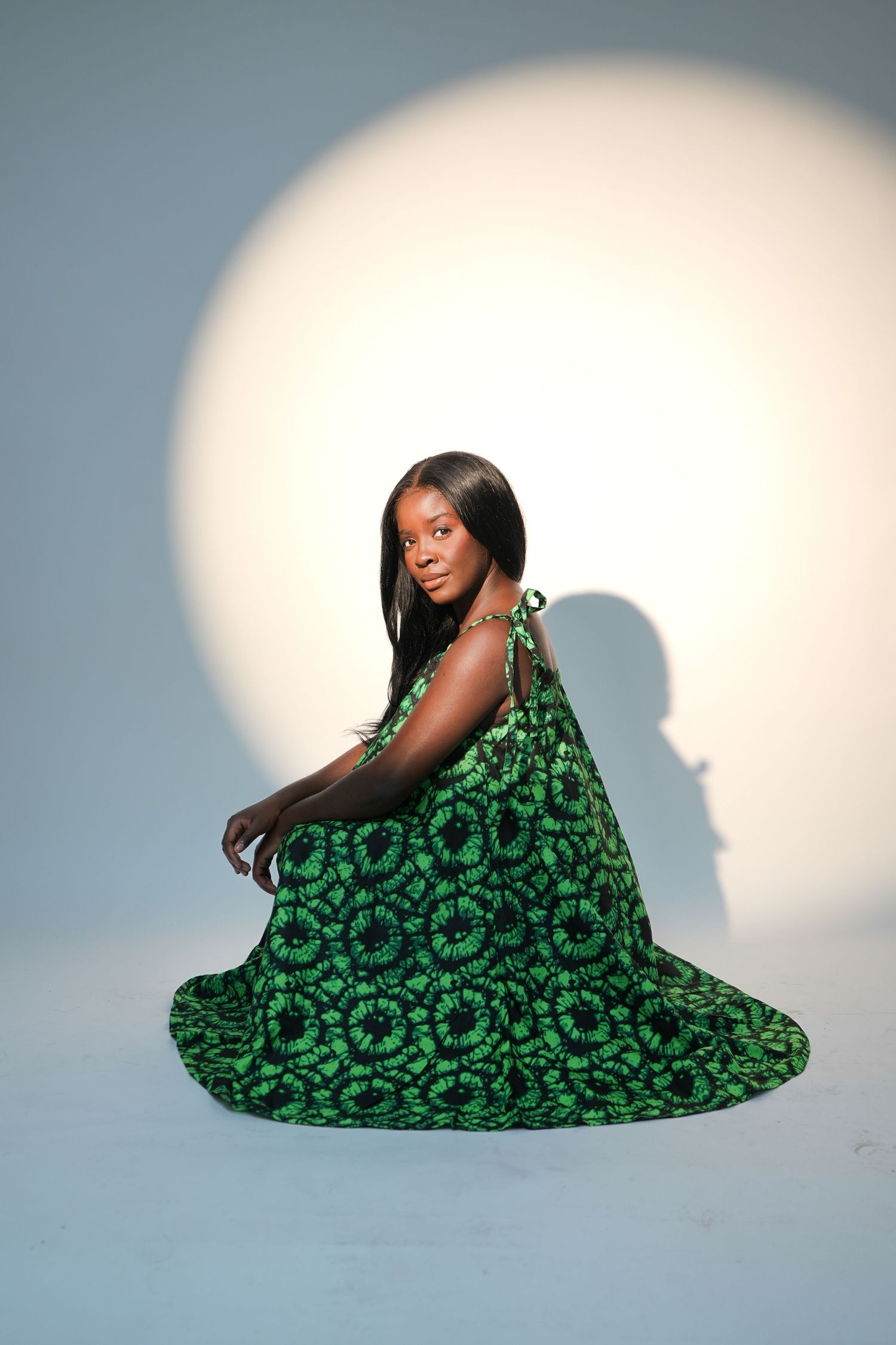 Woman in green African print dress sitting gracefully against a spotlight background in a studio photo.