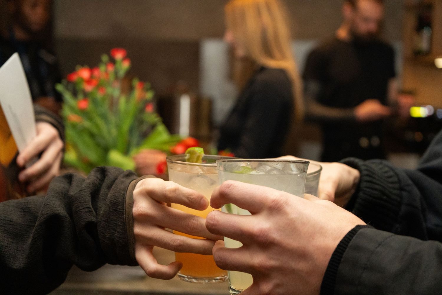 Two people clinking cocktail glasses at a bar, with flowers and guests in the background.