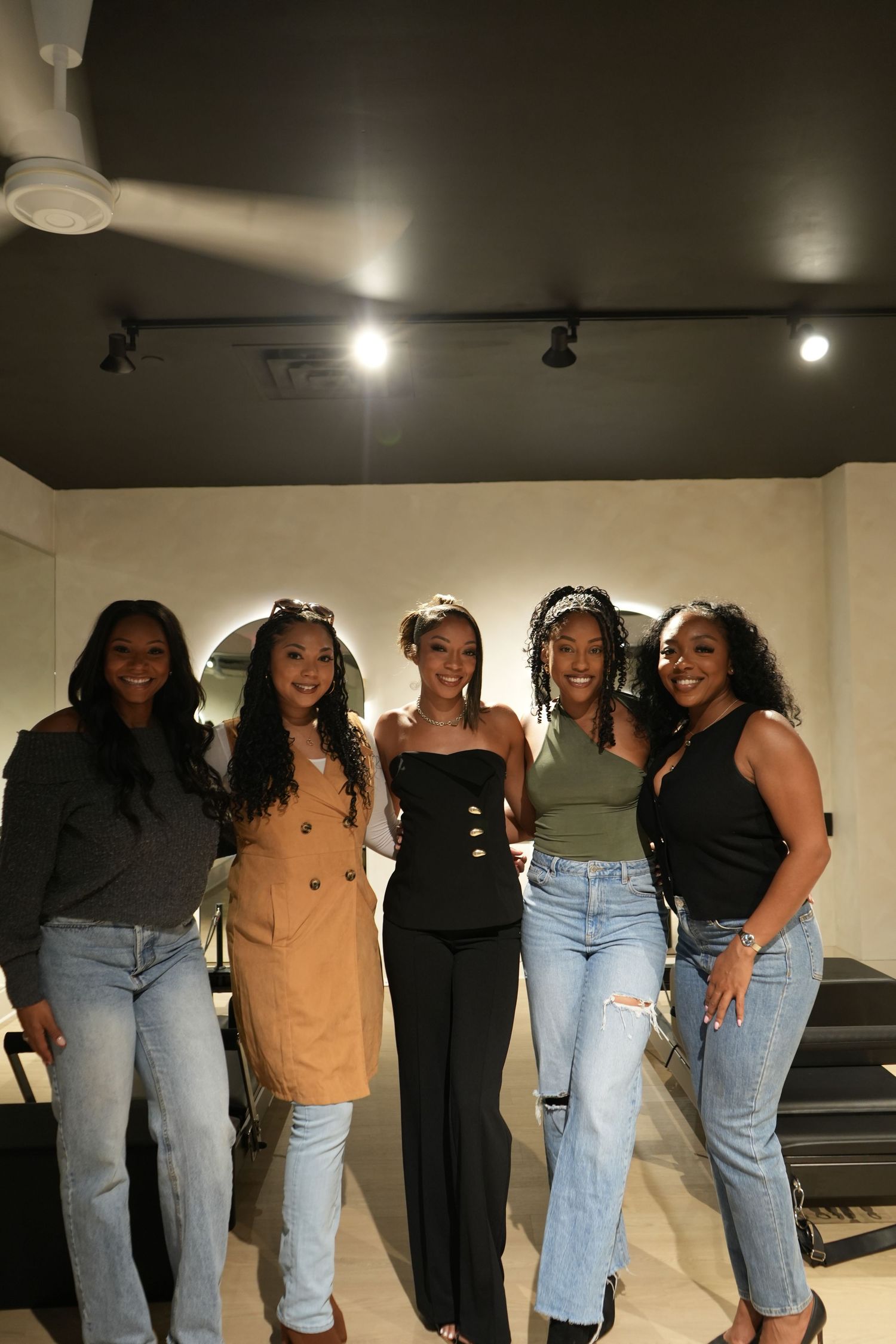 Five women smiling and posing together indoors under track lighting, wearing casual and stylish outfits.