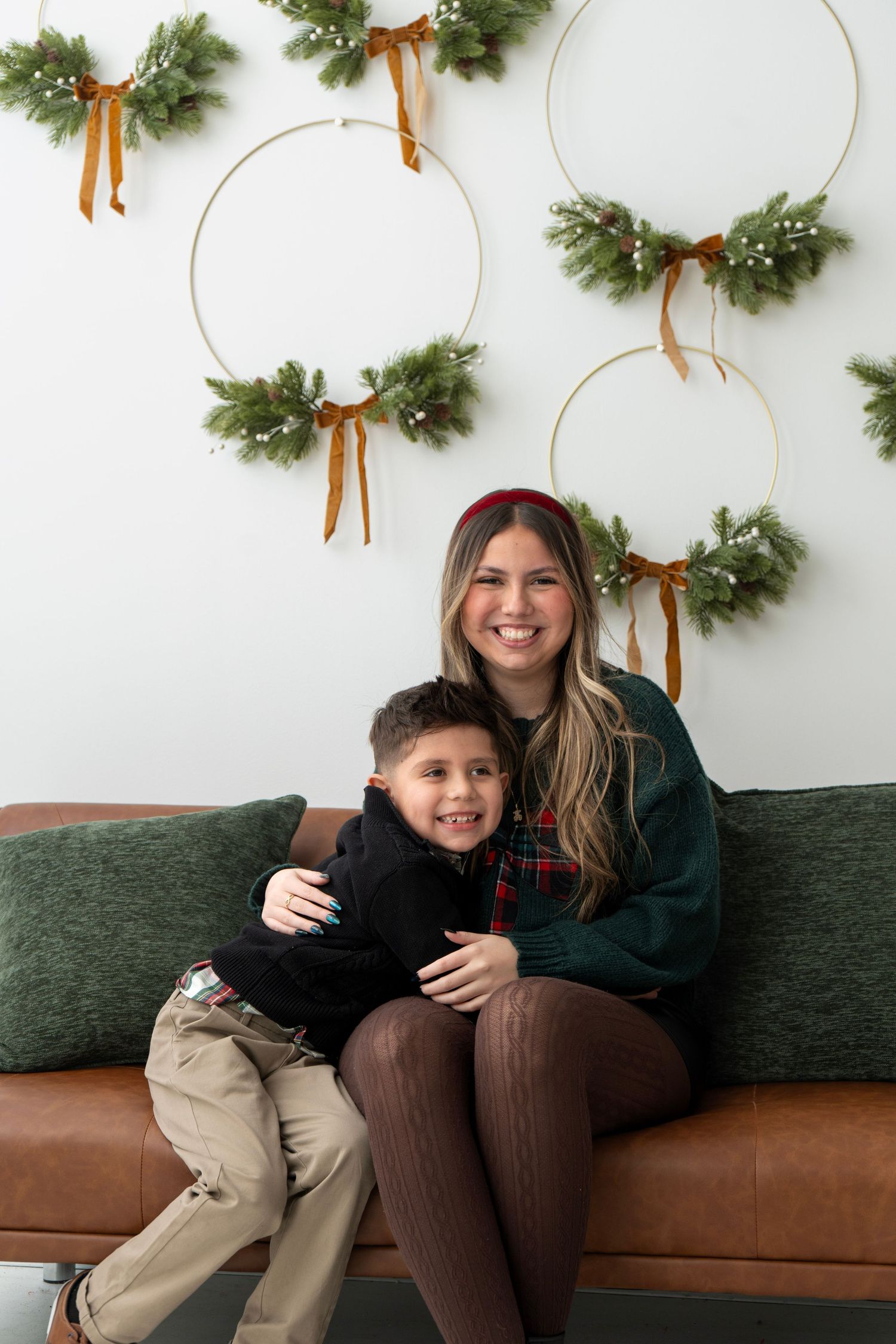 A woman and young boy smile together on a brown leather sofa with Christmas wreaths on the wall behind them.
