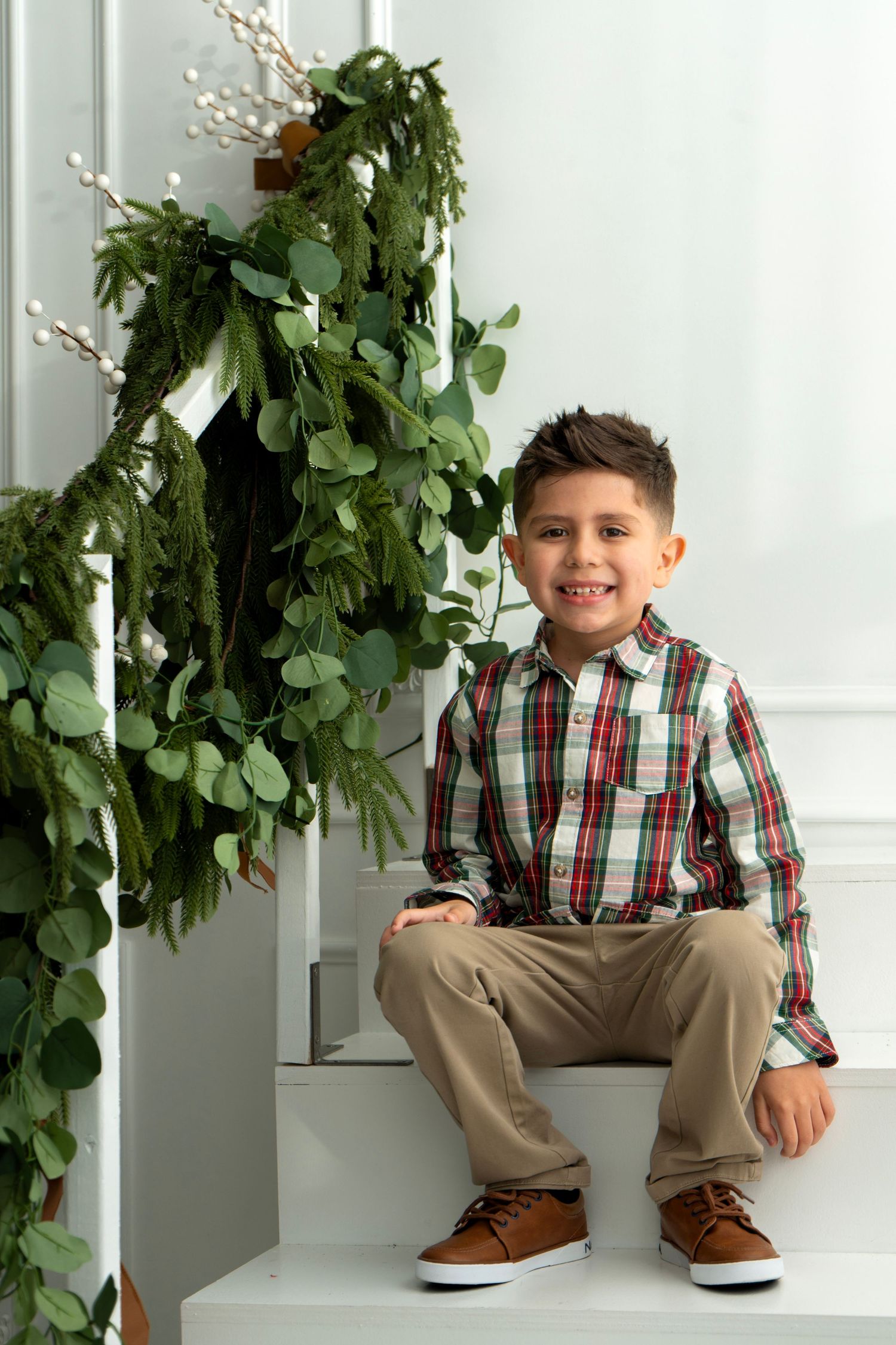 Smiling boy in plaid shirt and khakis sits on white stairs beside festive green holiday garland decoration.