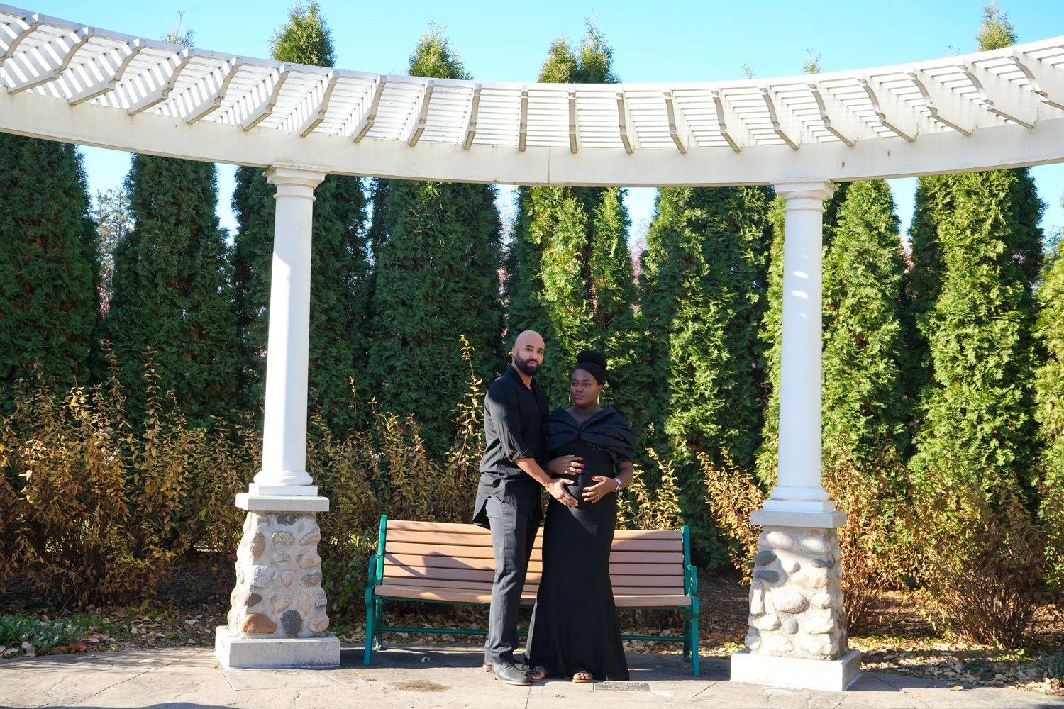 Two people dressed in black posing under a white pergola surrounded by lush green hedges and a wooden bench.