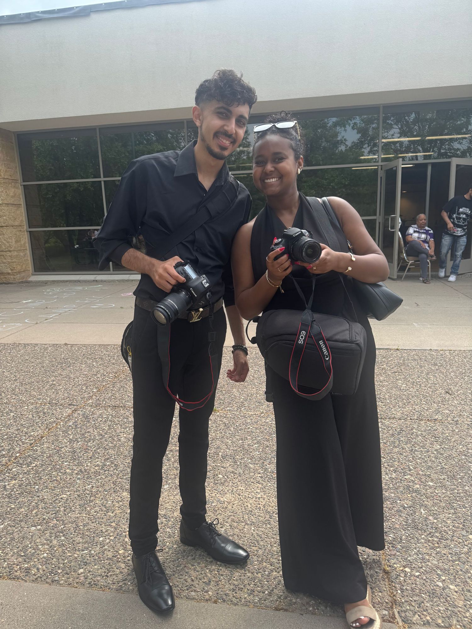 Two photographers pose with their cameras in a playful stance outside a building.