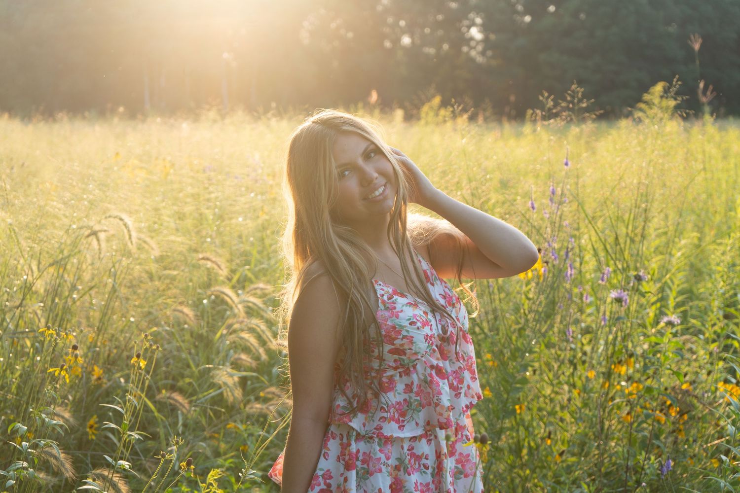 Young blonde woman in floral dress standing in sunlit meadow with wildflowers, golden hour light glowing behind her.
