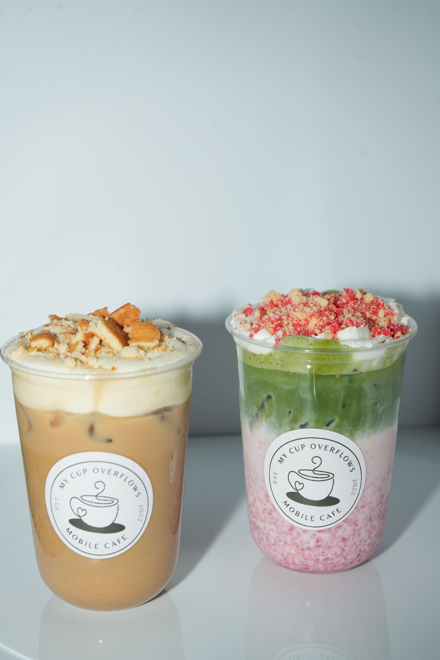 Two iced drinks from My Cup Overflows Mobile Cafe, one coffee and one matcha strawberry, topped with whipped cream and crumbles.