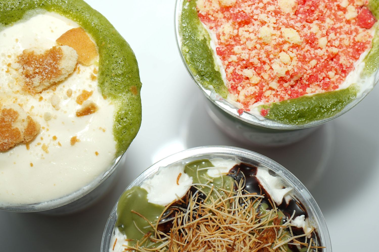 Three colorful dessert cups with green smoothie bases topped with cream, red crumbles, and noodle garnishes.