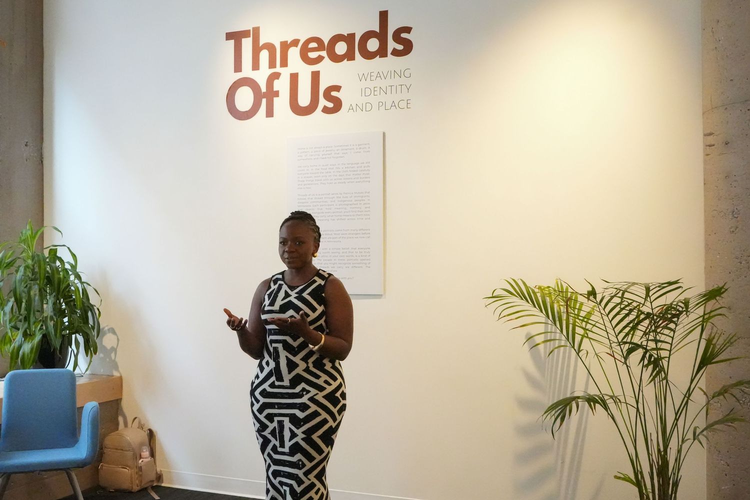 Woman in black and white dress stands before Threads Of Us exhibit sign with tropical plants nearby.