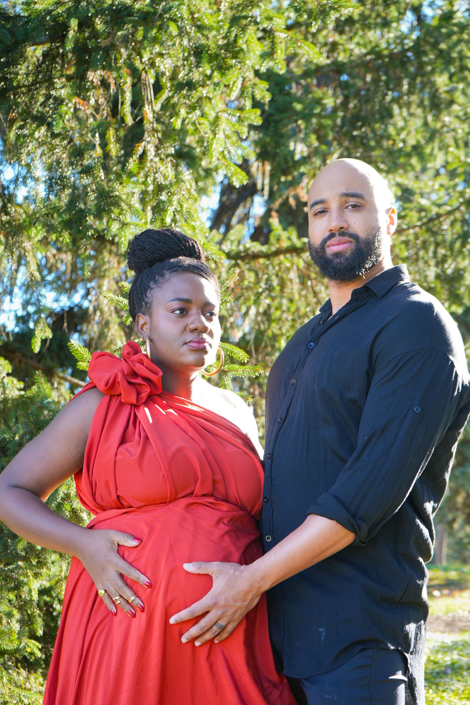 Pregnant woman in red dress and man in black shirt pose together outdoors near green trees for maternity photo.