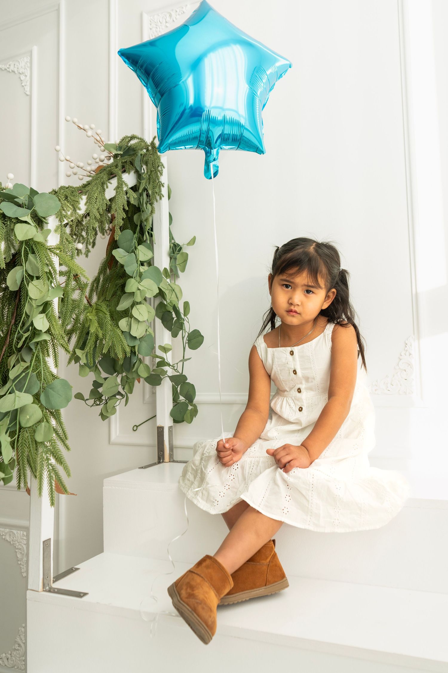 Young girl in white dress and brown boots sits on white ledge holding blue star balloon beside greenery.