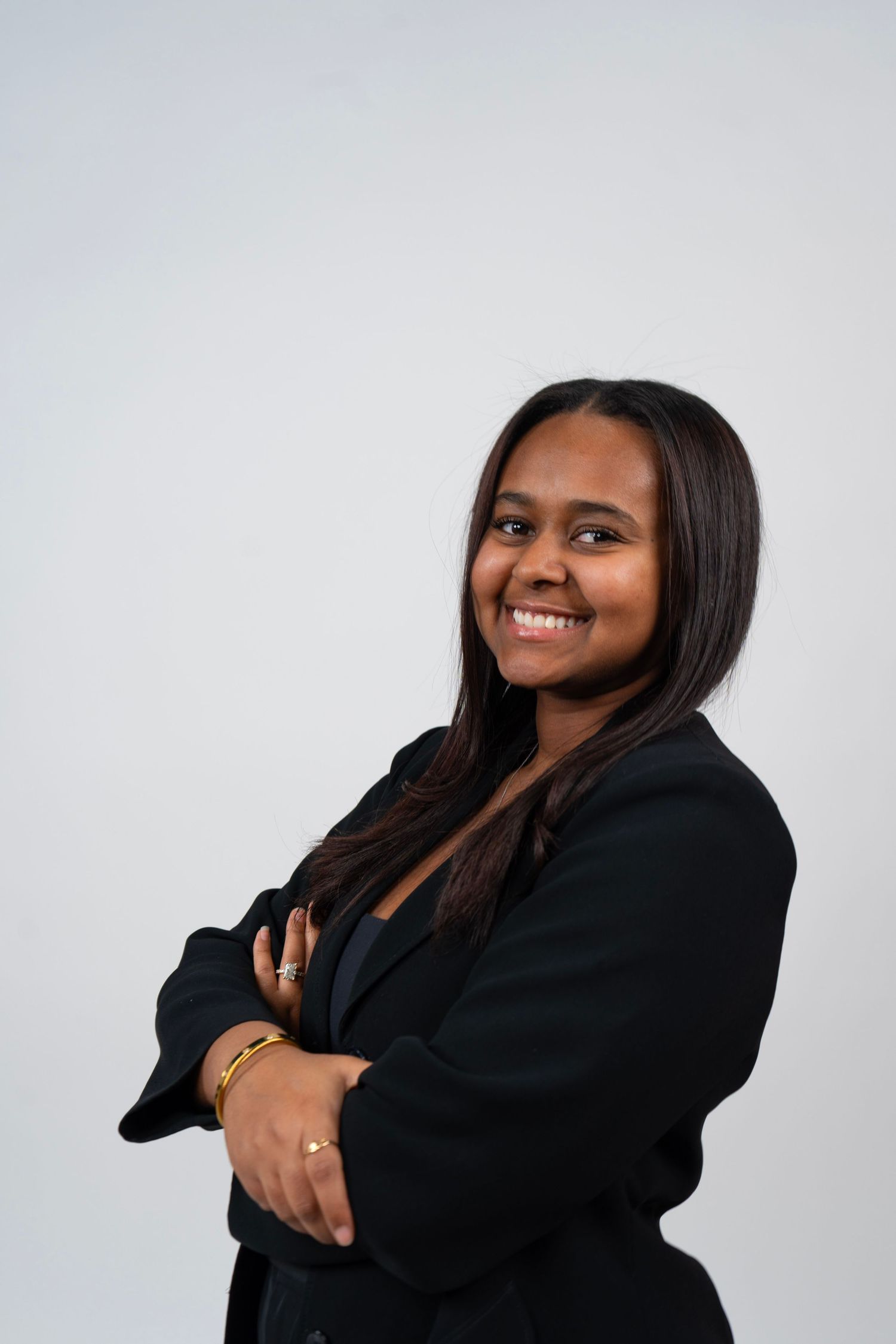 Smiling young woman in black blazer with arms crossed, wearing gold bracelet, posing confidently against gray background.