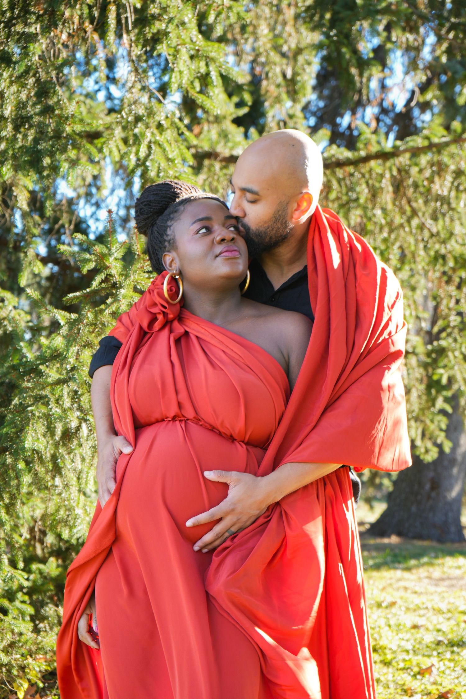 Pregnant couple in red fabric wrap embracing outdoors among evergreen trees in a maternity photo shoot.