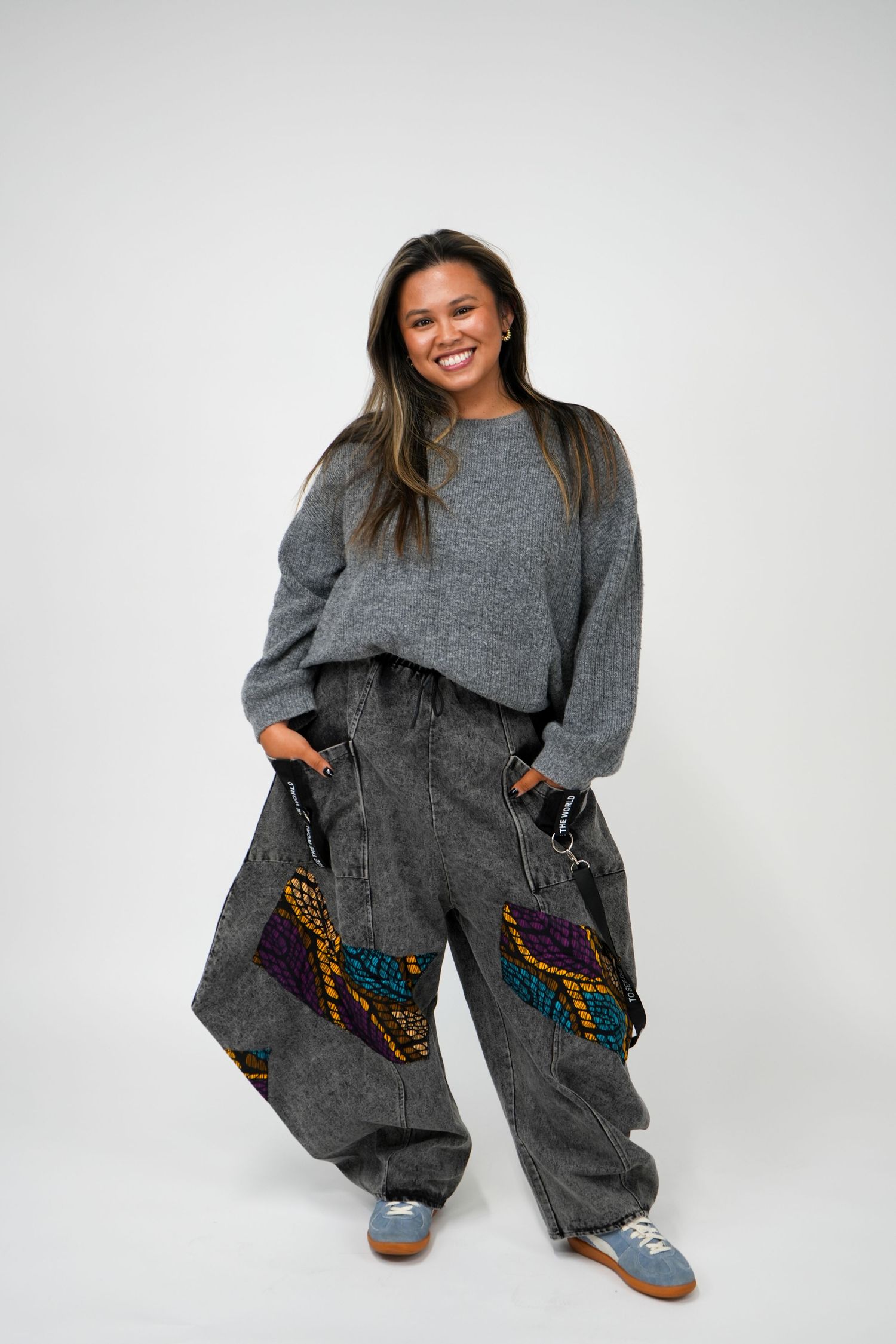 Woman smiling in gray oversized sweater and wide-leg acid wash pants with colorful patchwork detail.