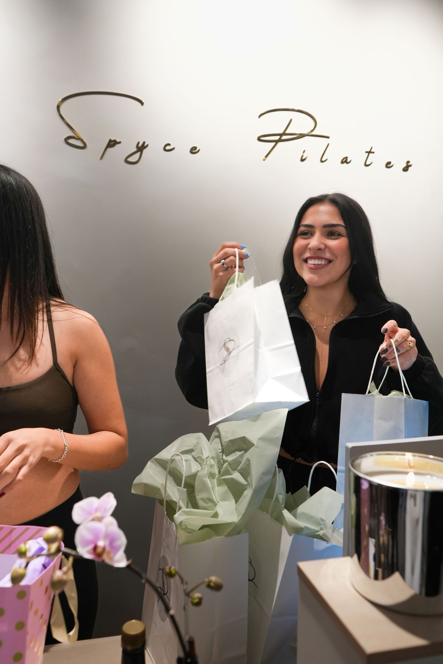 Two women smiling and holding gift bags at Spyce Pilates studio, surrounded by elegant packaging and candles.