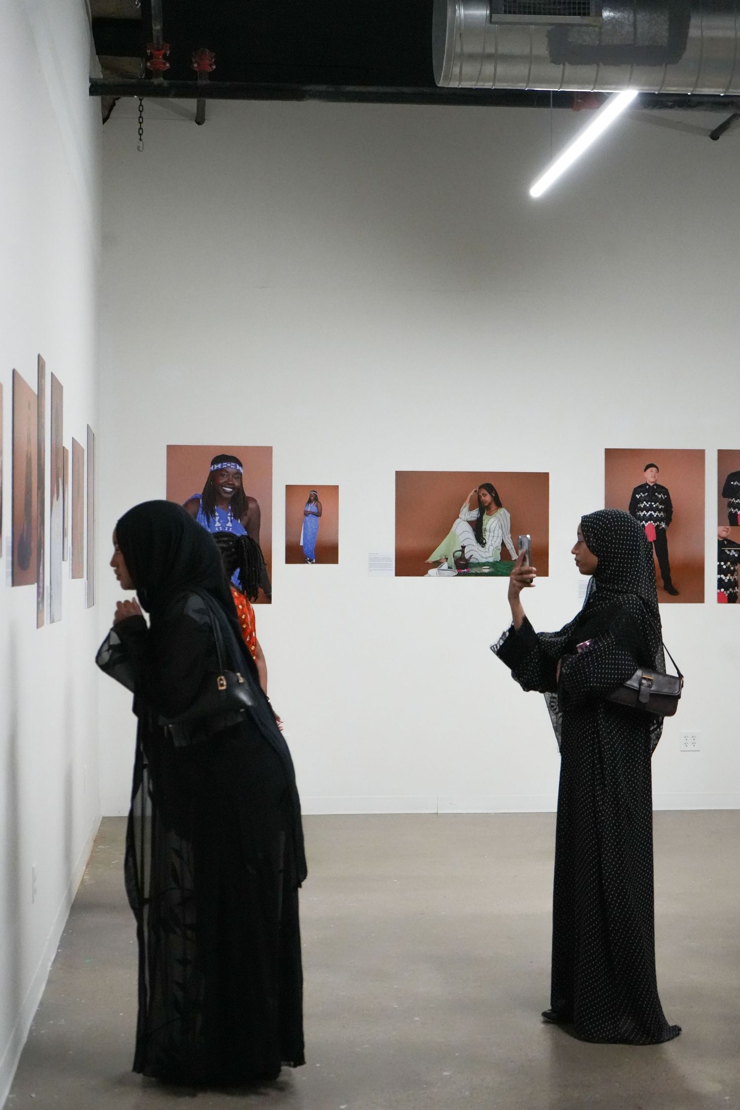 Two women in black abayas view fashion photography prints displayed on white gallery walls.