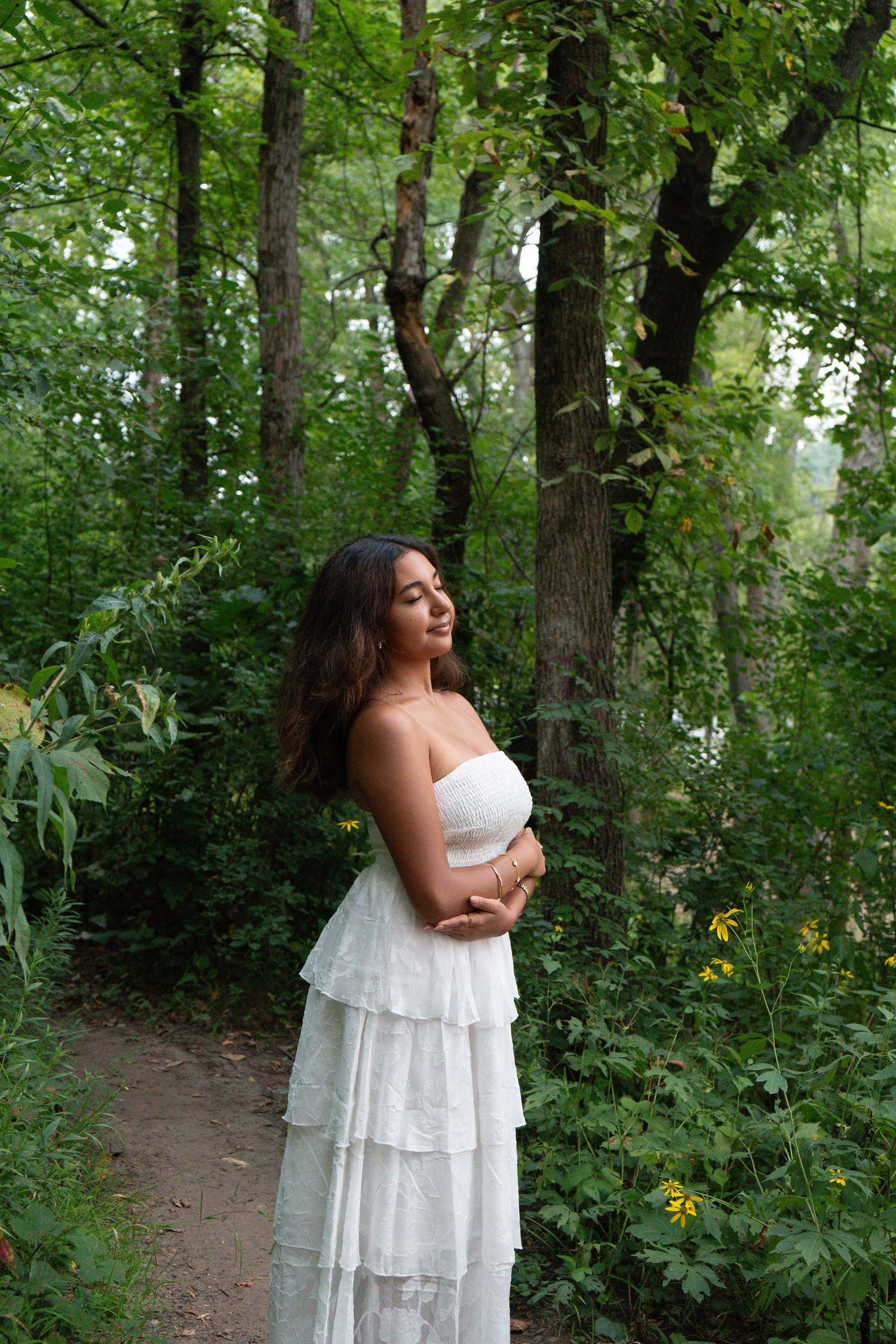 A woman in a white strapless ruffled dress stands gracefully on a forest path surrounded by lush green trees.