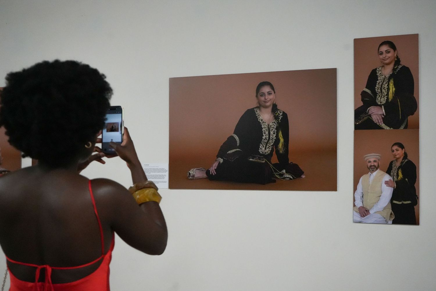 Woman photographs gallery wall displaying portraits of South Asian individuals in traditional attire at an art exhibition.