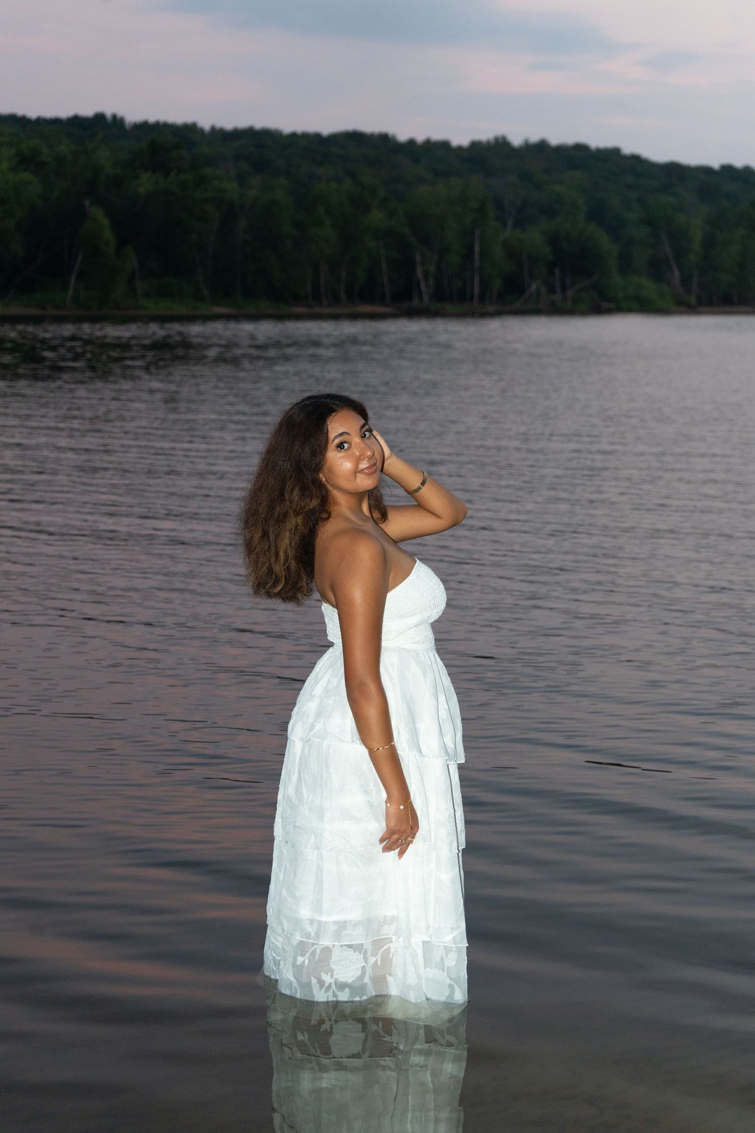 Young woman in white dress standing in a calm lake at dusk, with lush green forest in the background.